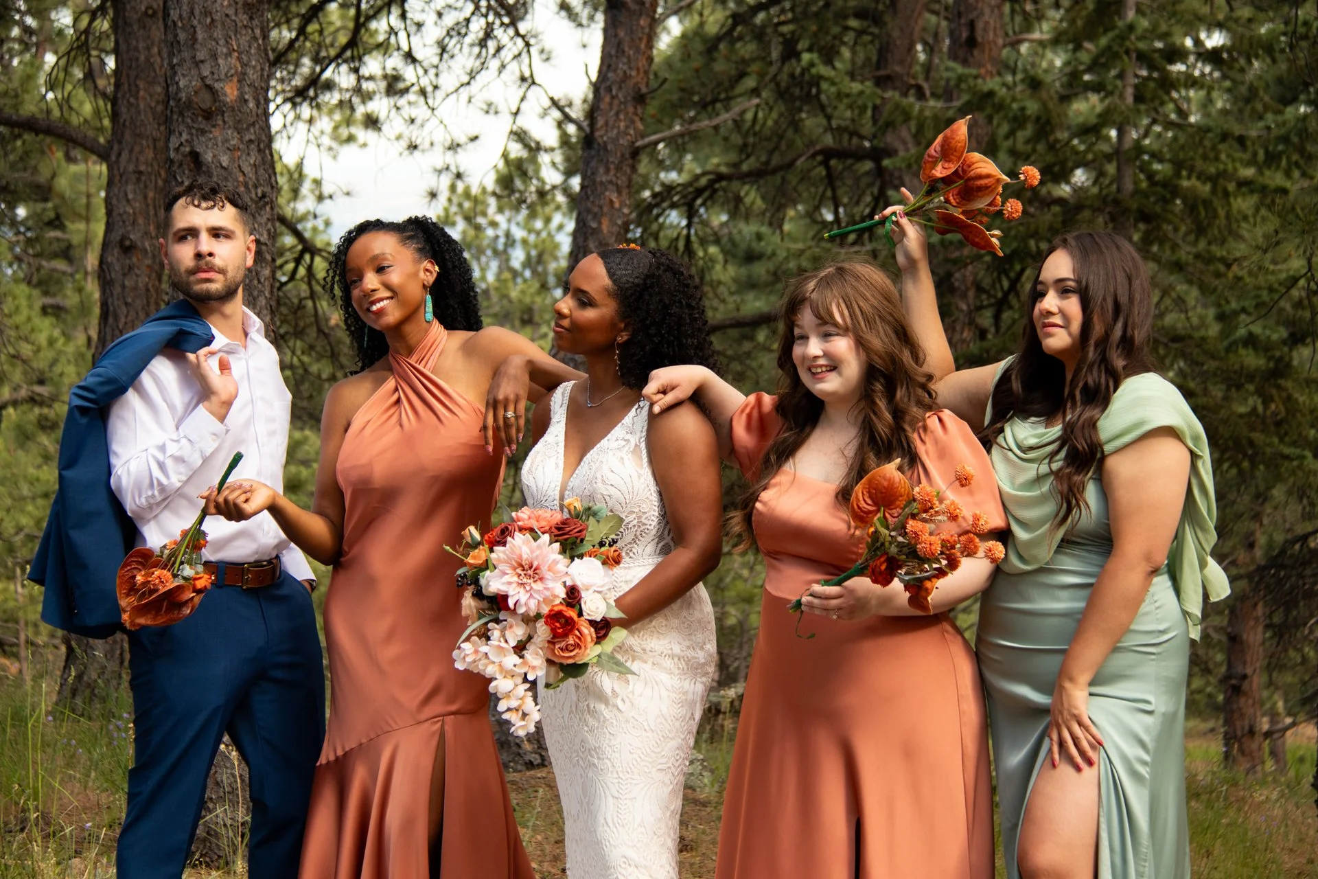A group of six diverse women and one man posing outdoors in a forest, dressed in colorful formal dresses and suits, holding bouquets of flowers, with some smiling playfully.