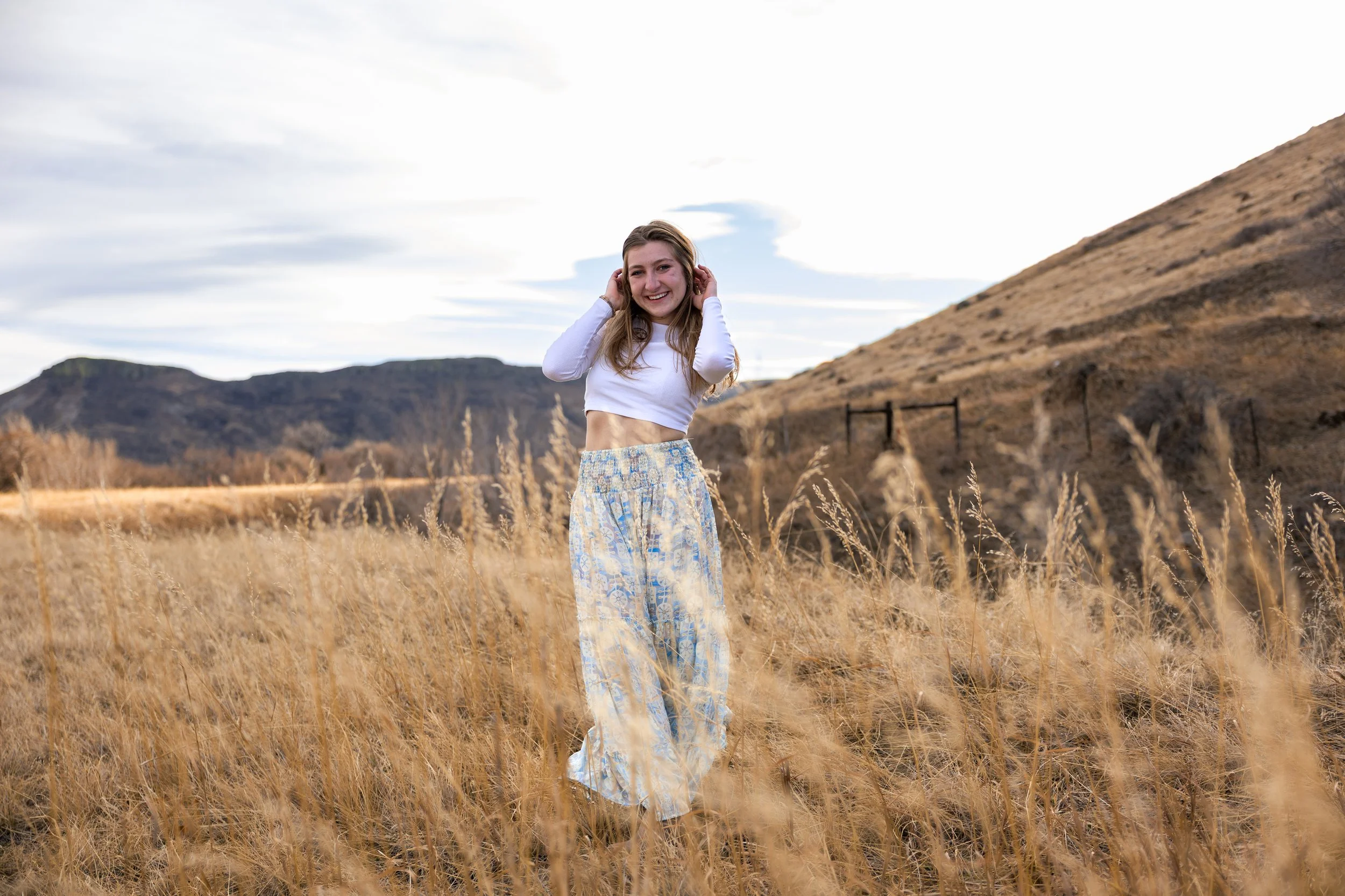 A young woman standing in a field of tall, golden grass with hills and mountains in the background, smiling and adjusting her hair.