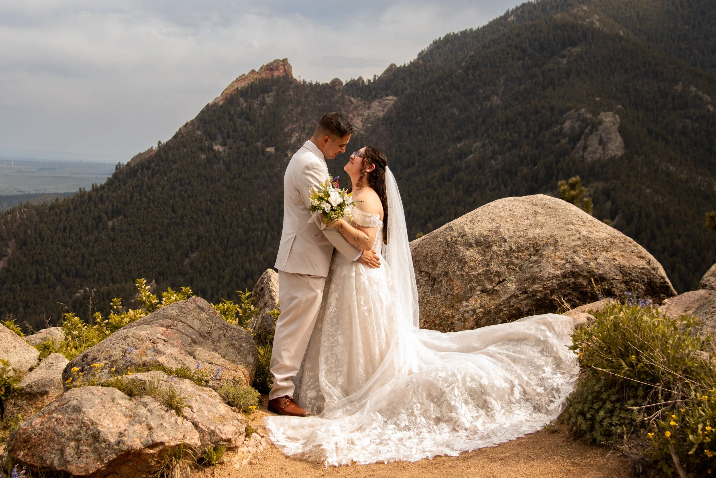 A newlywed couple stands on a rocky hilltop with mountains in the background. The bride is in a white lace wedding gown with a long train and veil, holding a bouquet of flowers. The groom is in a beige suit. They are gazing at each other lovingly.