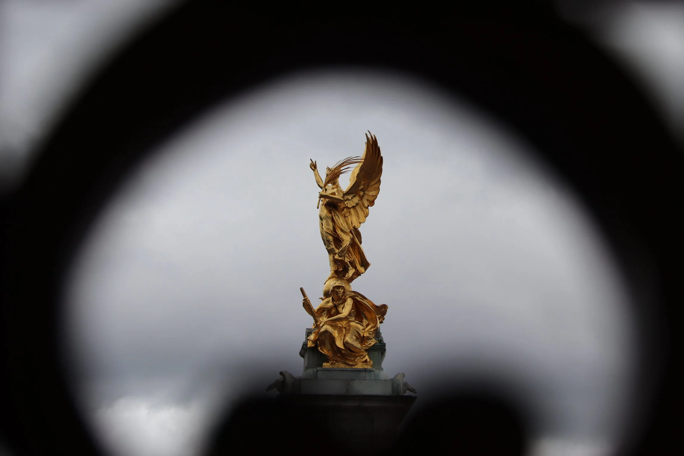 Golden monument of figures with wings, topped by a winged female figure, viewed through a circular frame with cloudy sky background.