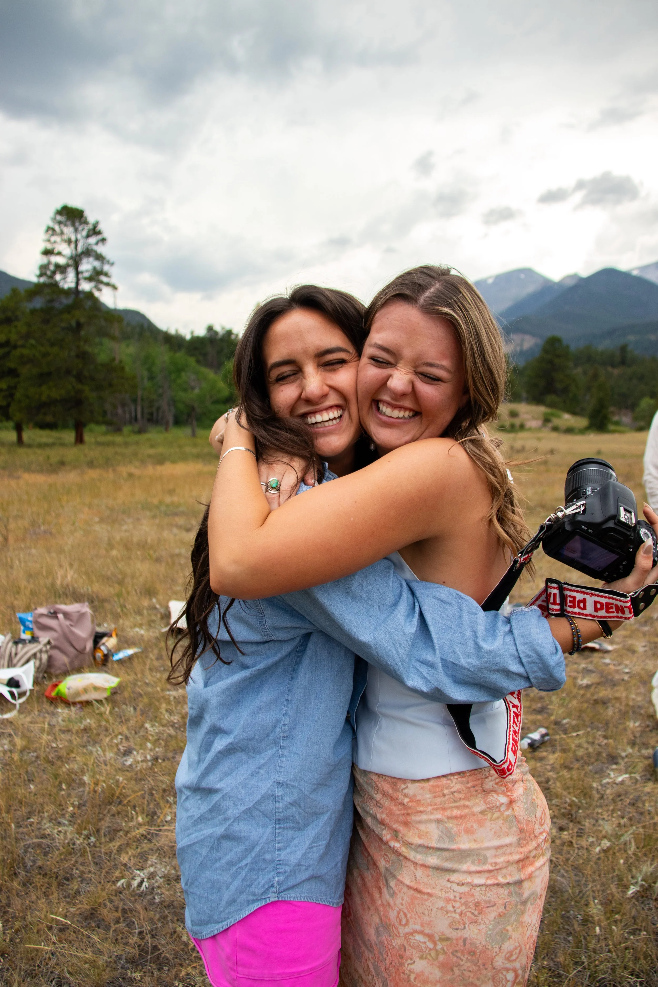 Two young women hugging and smiling outdoors in a grassy field with trees and mountains in the background, one holding a camera.
