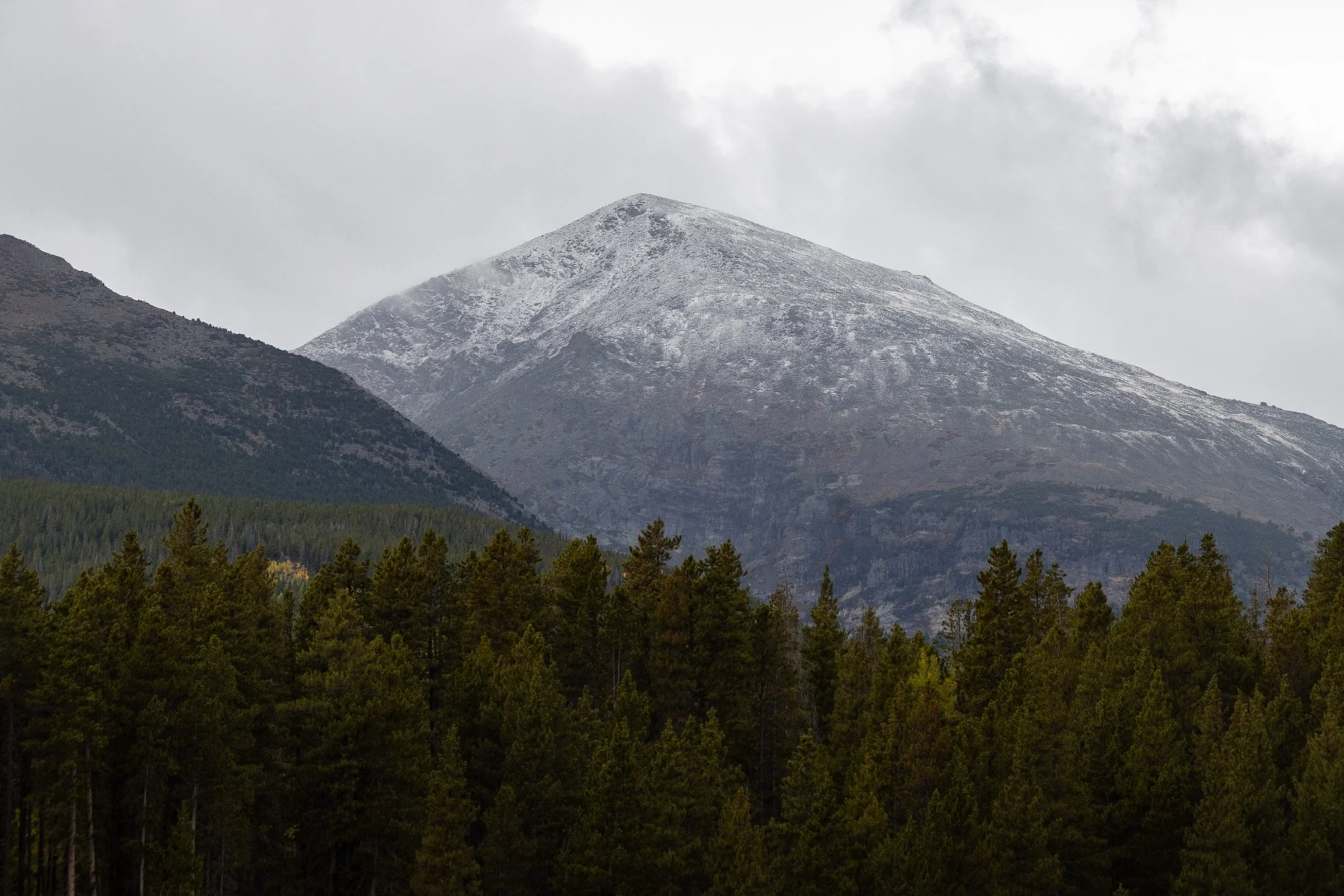 Mountain with some snow on top, over a forest of pine trees.