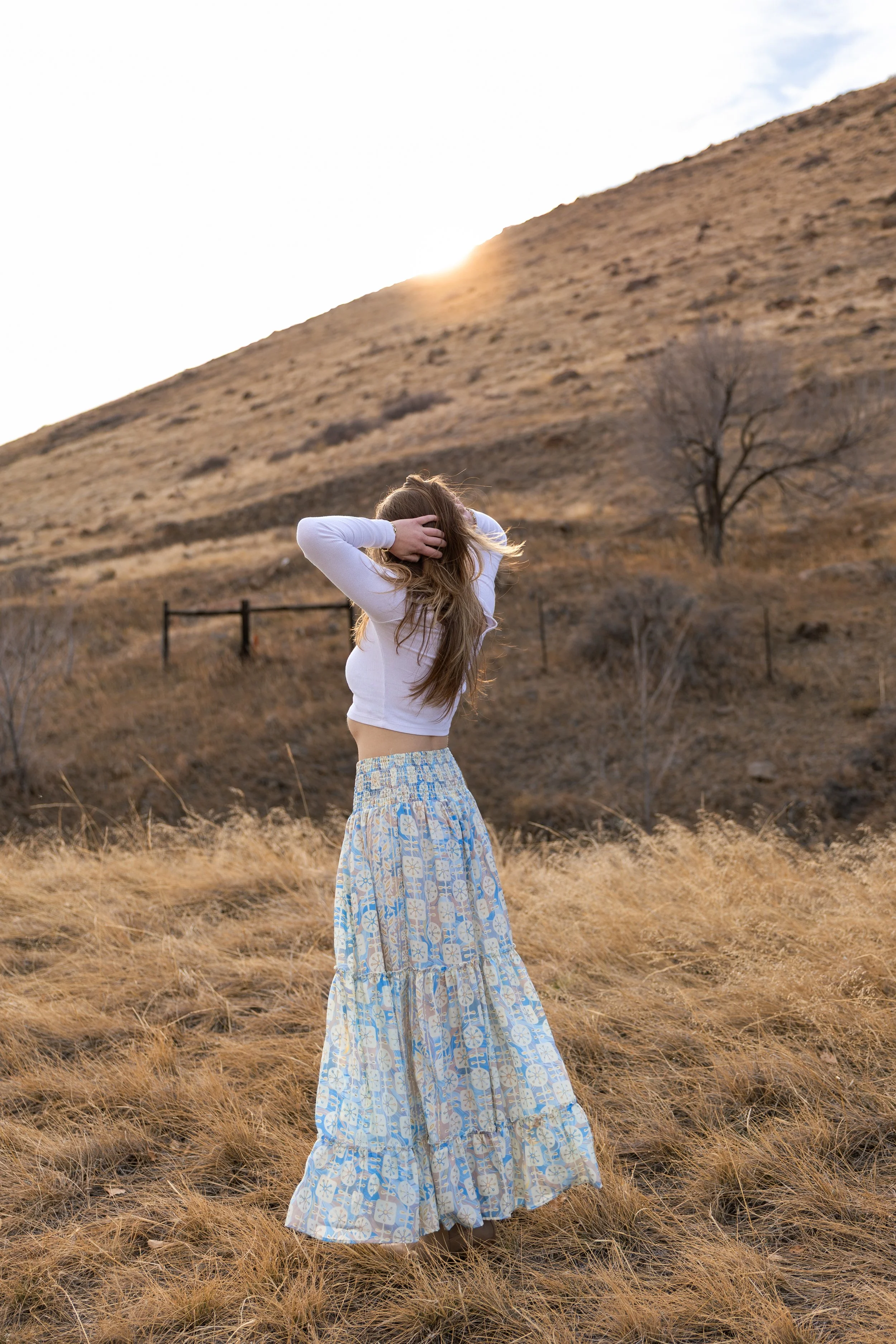 A woman standing in a dry field of tall grass with a hillside and a few trees in the background, during sunset.