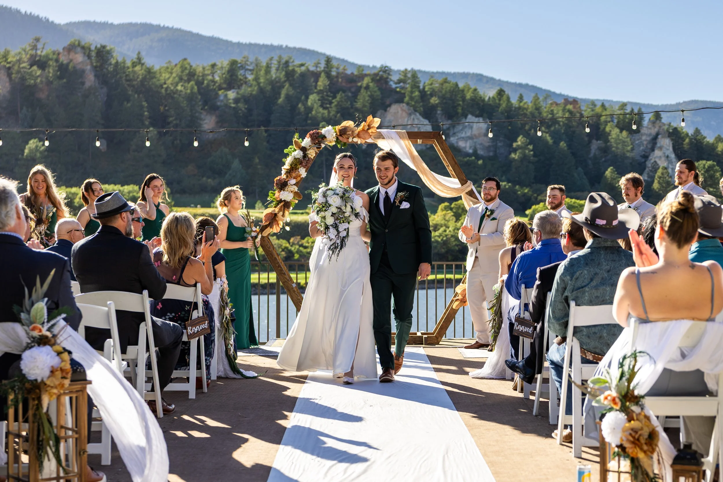 A bride and groom walk down the aisle at their outdoor wedding ceremony in scenic mountains, surrounded by seated guests and wedding party, with a decorative floral arch behind them