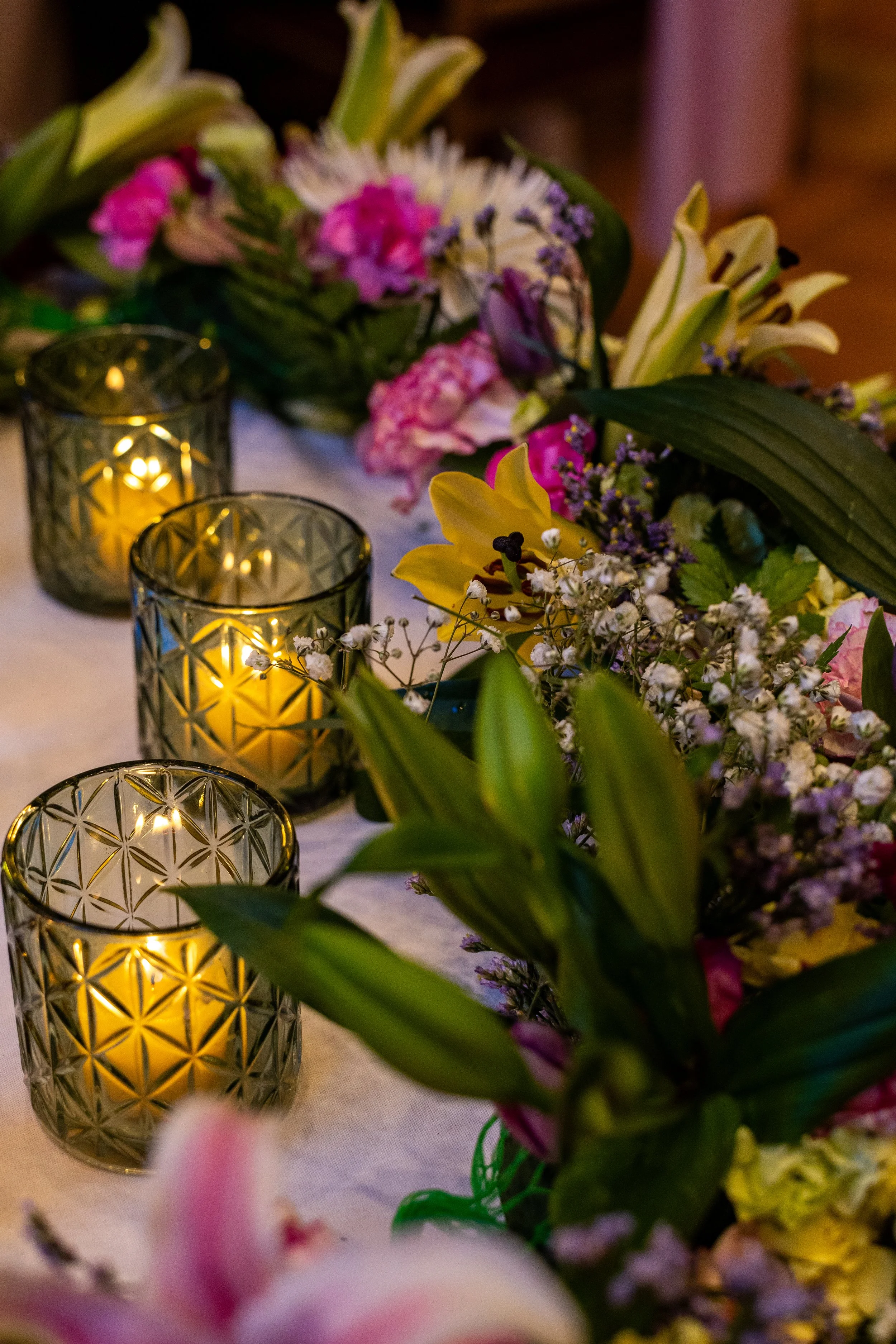 Vase filled with colorful flowers, including lilies and pink flowers, surrounded by three lit candles in decorative glass holders on a table.