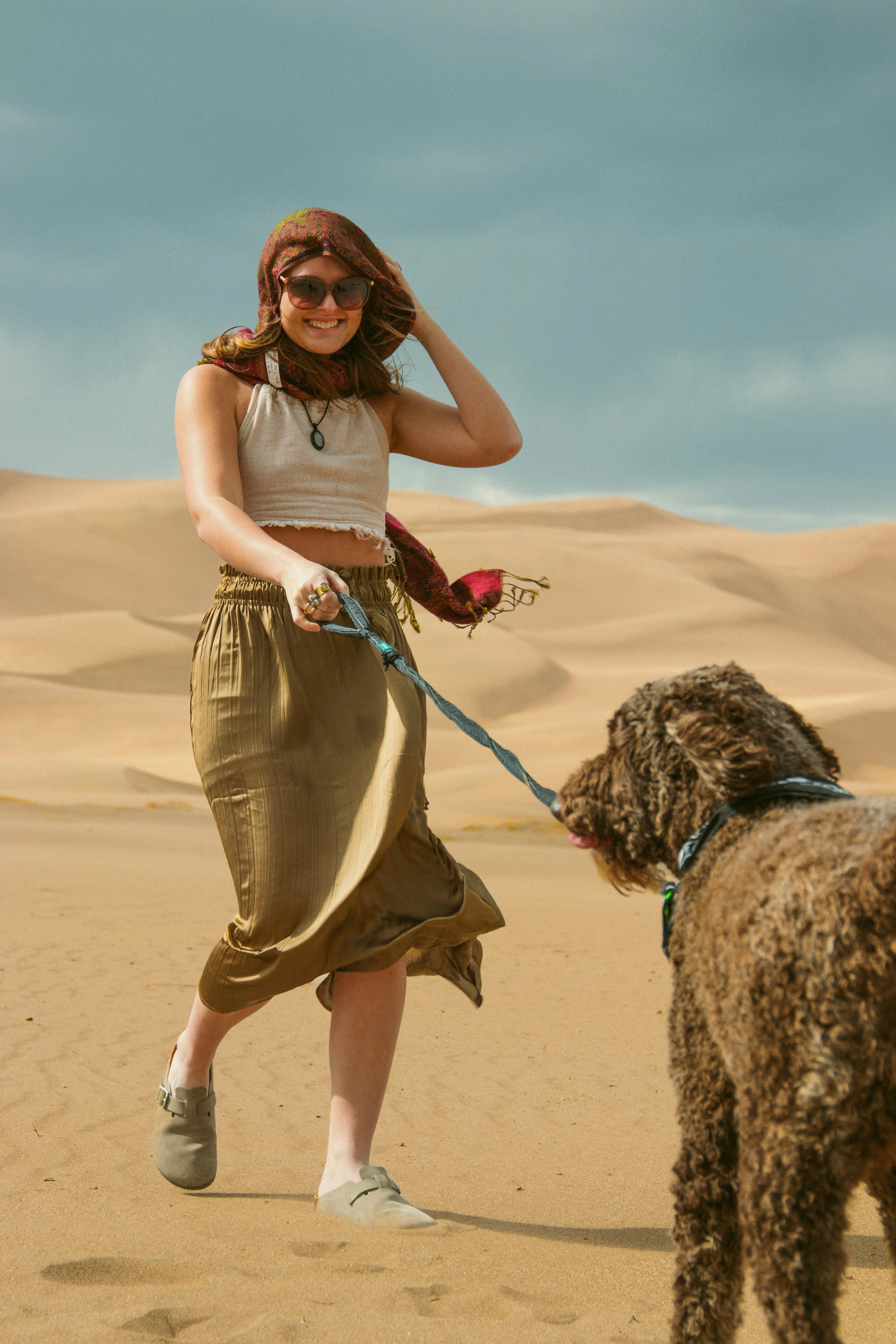 A woman in sunglasses and a beige crop top walking a curly-haired dog in the Sand Dunes National Park.