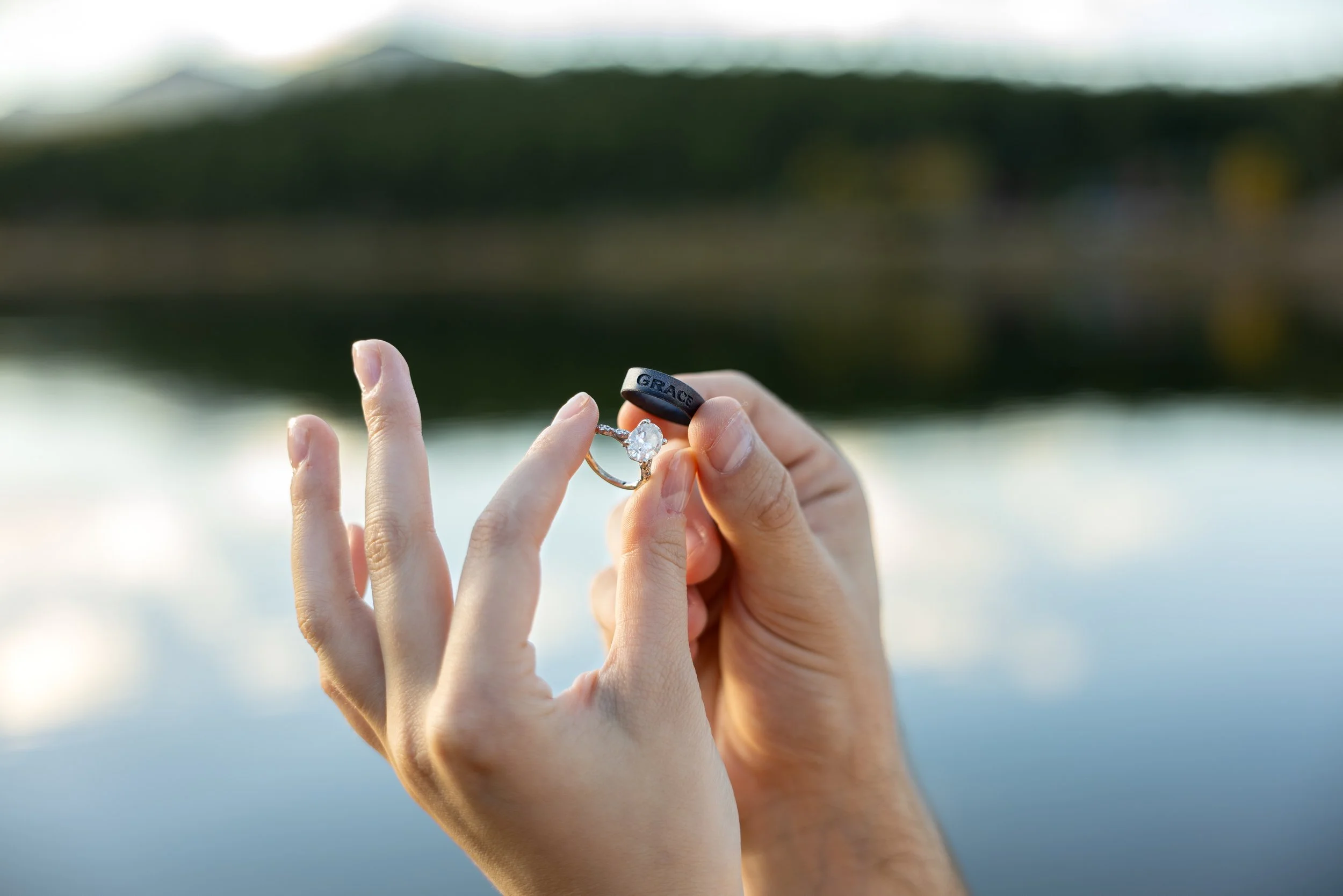 A pair of hands holding an engagement ring with a diamond and a black protective ring engraved with the word 'GRACE,' against a blurred background of water and greenery.