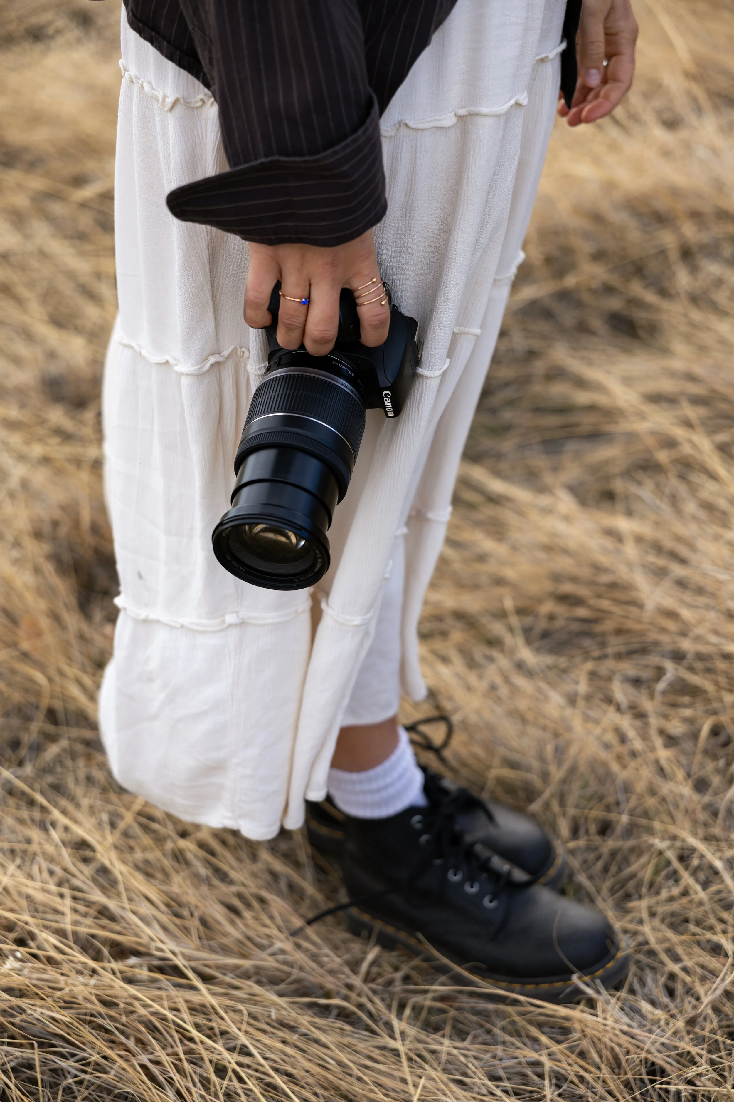 Person holding a camera with a long lens, standing on dry grass, wearing black shoes, white socks, a white skirt, and a black top with a striped arm sleeve.