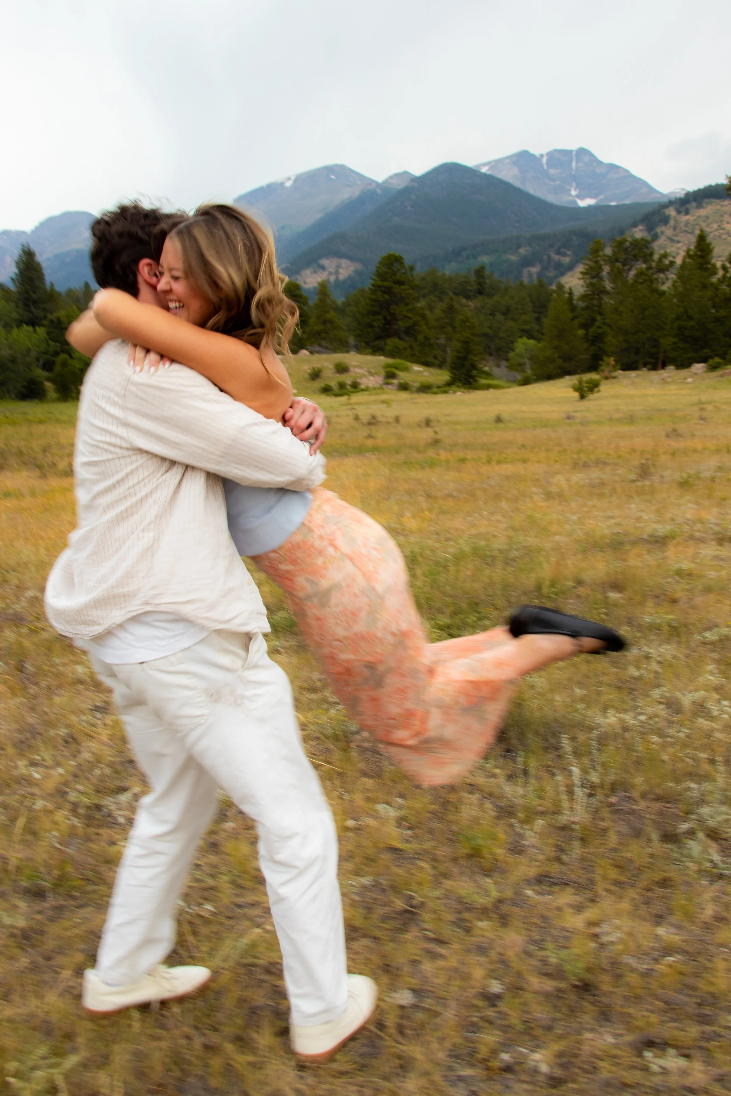 A man lifting and spinning a woman in a grassy field with mountains and trees in the background, both smiling and enjoying the moment.