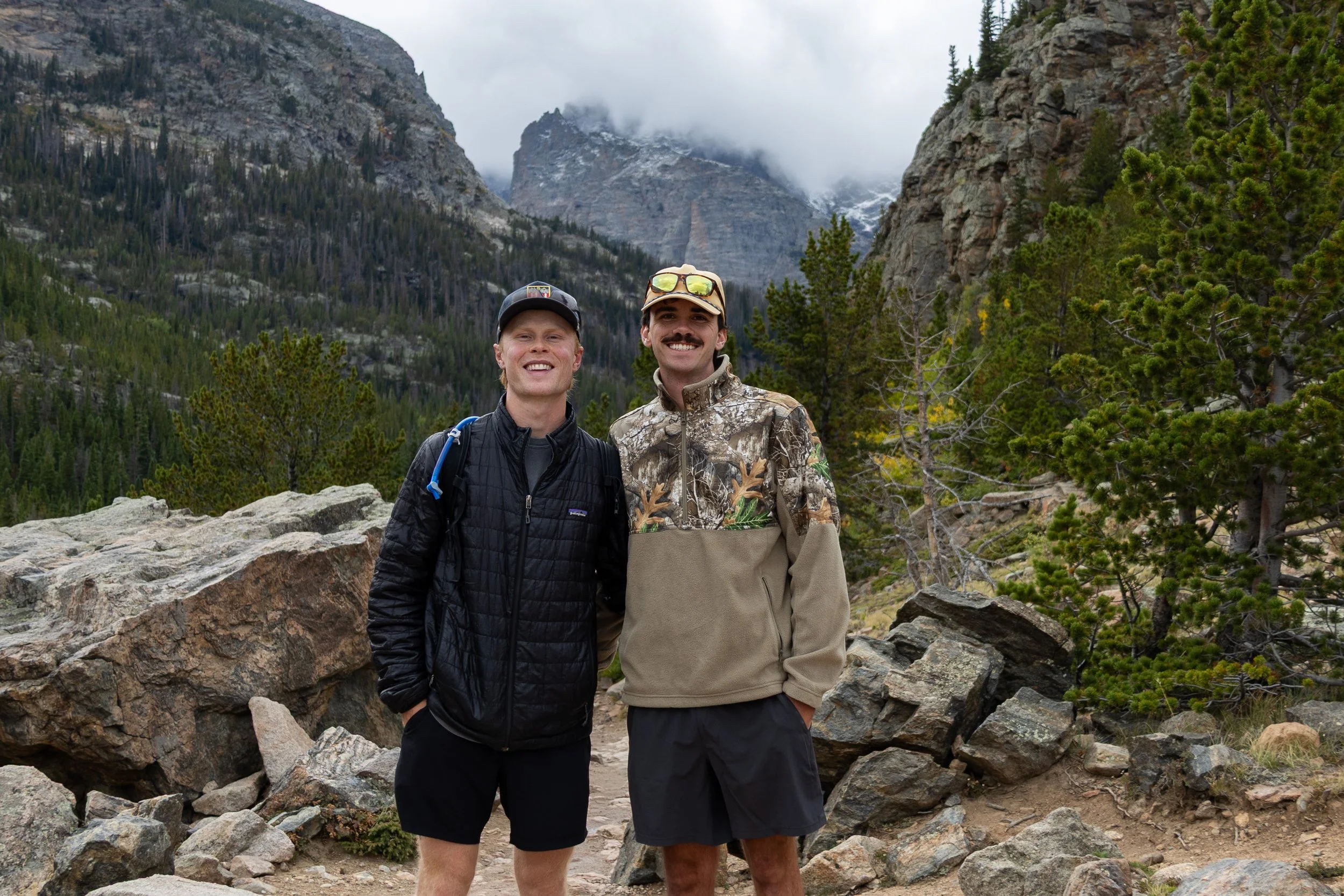 Two men smiling outdoors in Rocky Mountain National Park with rocks and pine trees around them.