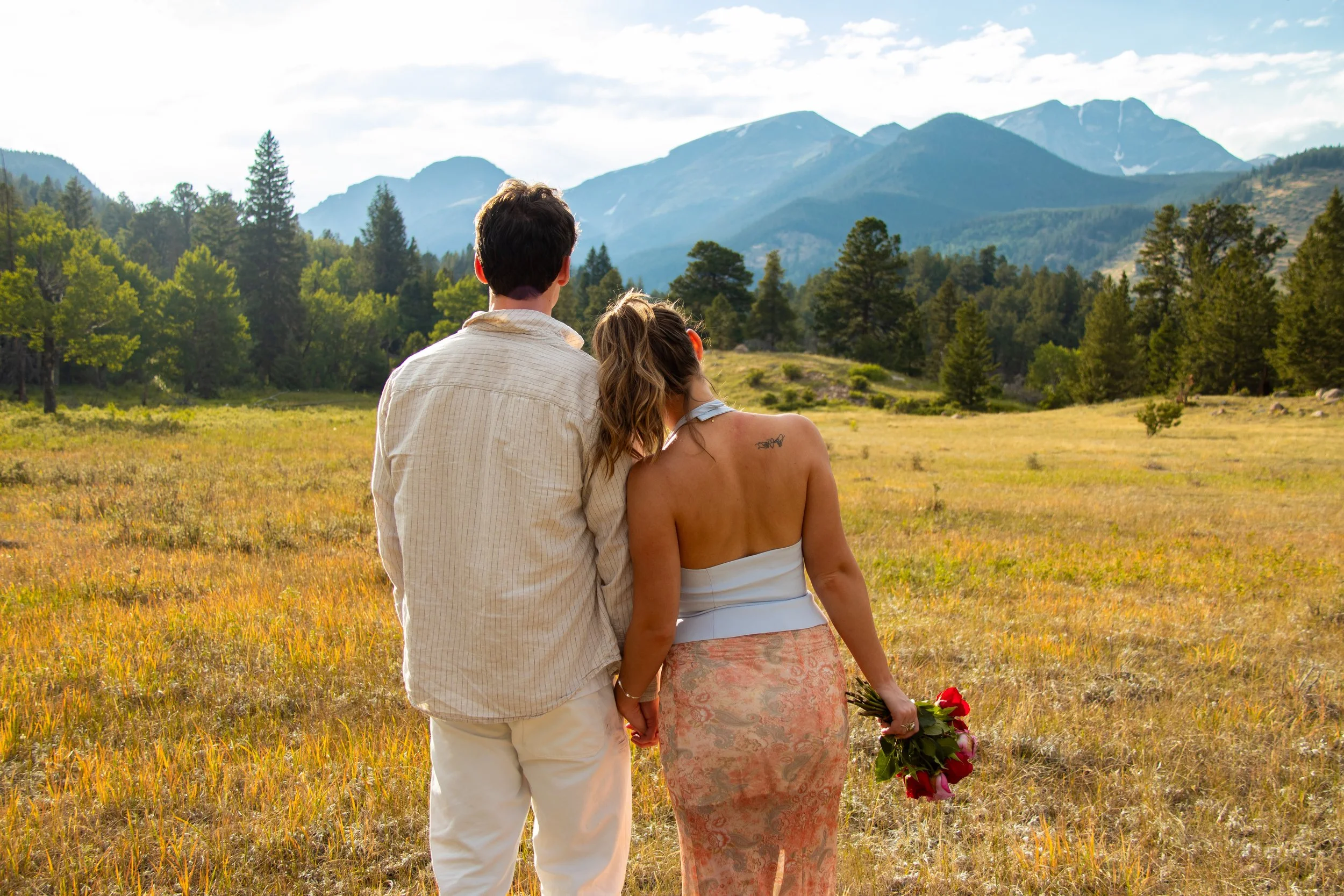 A couple holding hands in a field with mountains in the background. The woman is holding a bouquet of flowers.