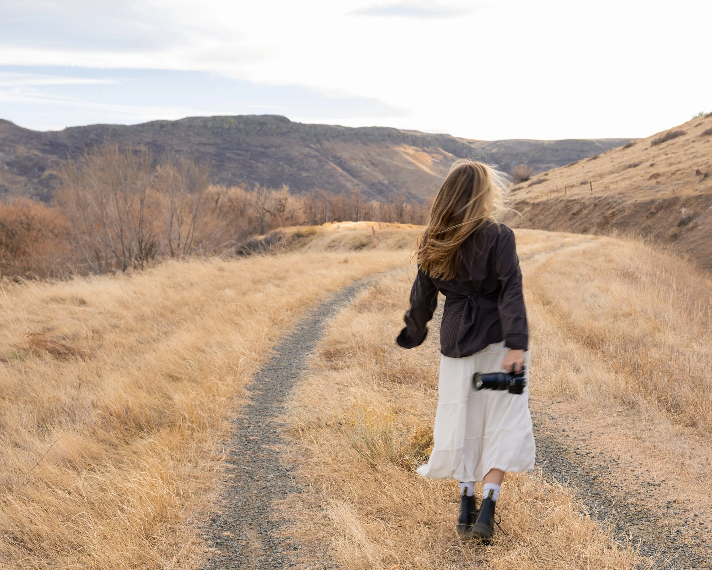 Woman walking on a dirt trail through a dry, grassy landscape with rolling hills in the background, holding a camera in her right hand, with wind blowing her hair.