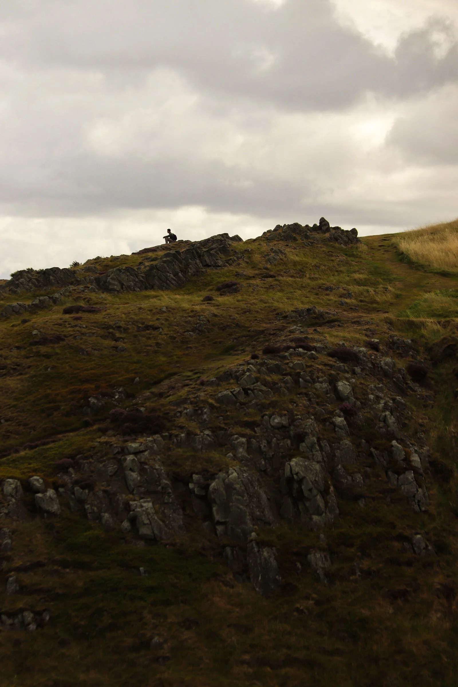 A person sitting on a rock formation on a sloped hillside under cloudy sky.