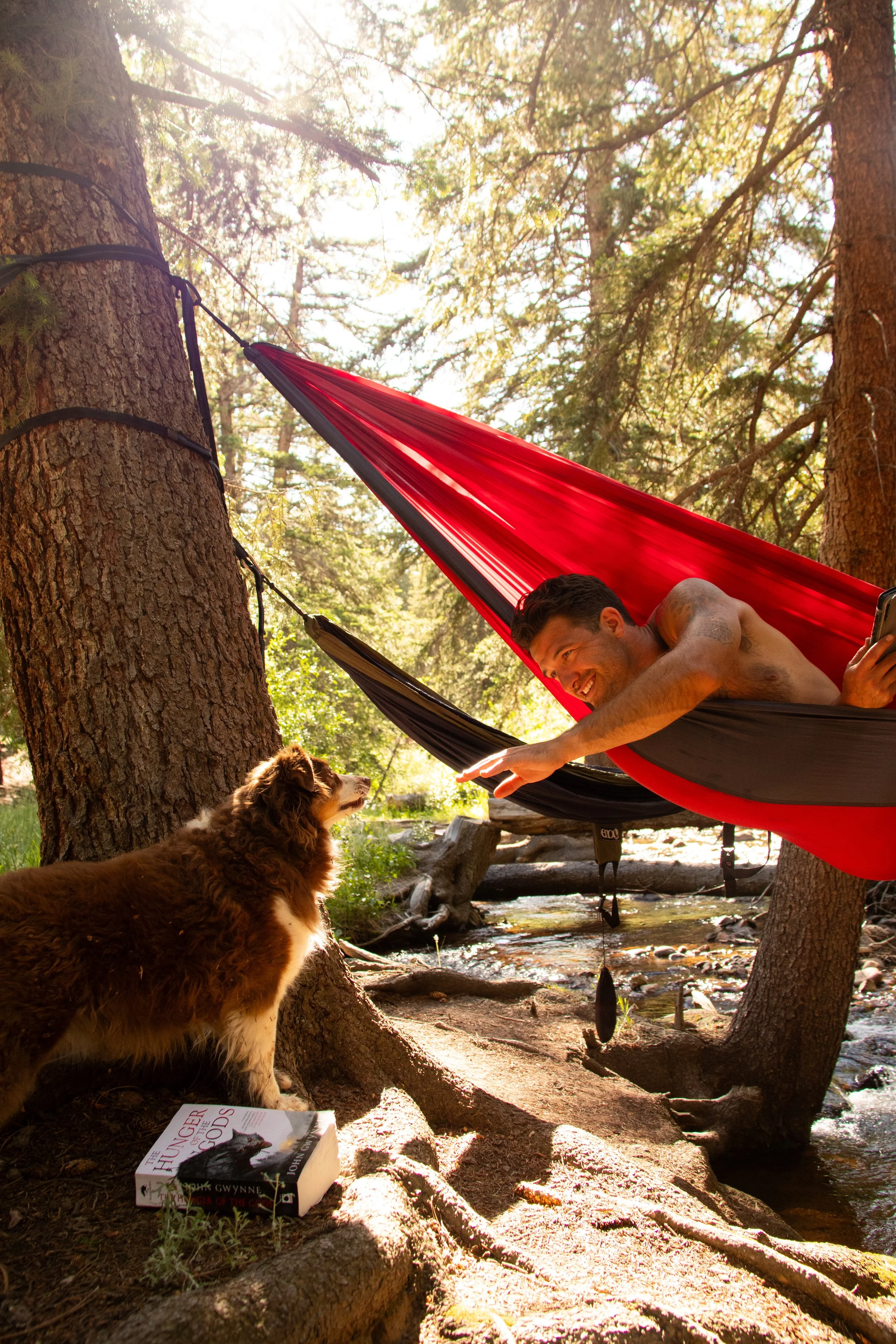 A man lying shirtless in a red and black hammock between trees in a wooded area, reaching out to a dog standing on the ground.