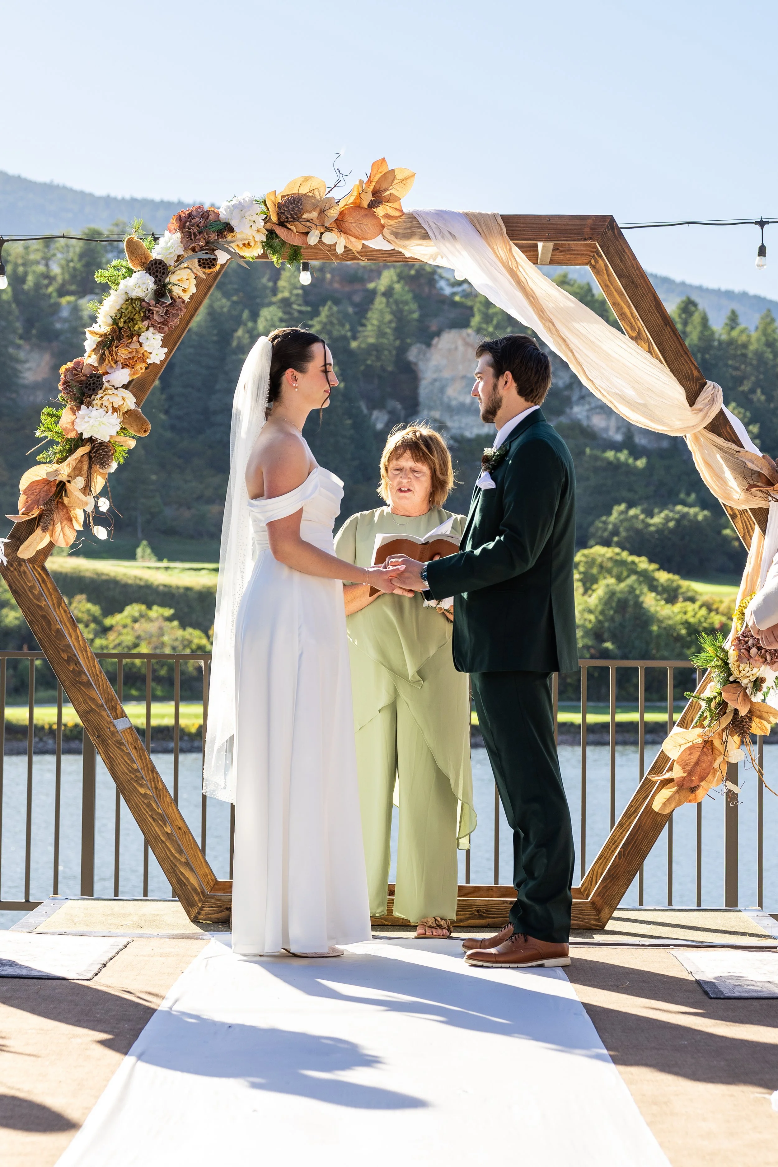 A bride and groom exchanging vows during an outdoor wedding ceremony, standing under a wooden arch decorated with flowers and draped fabric, with scenic mountains and trees in the background.