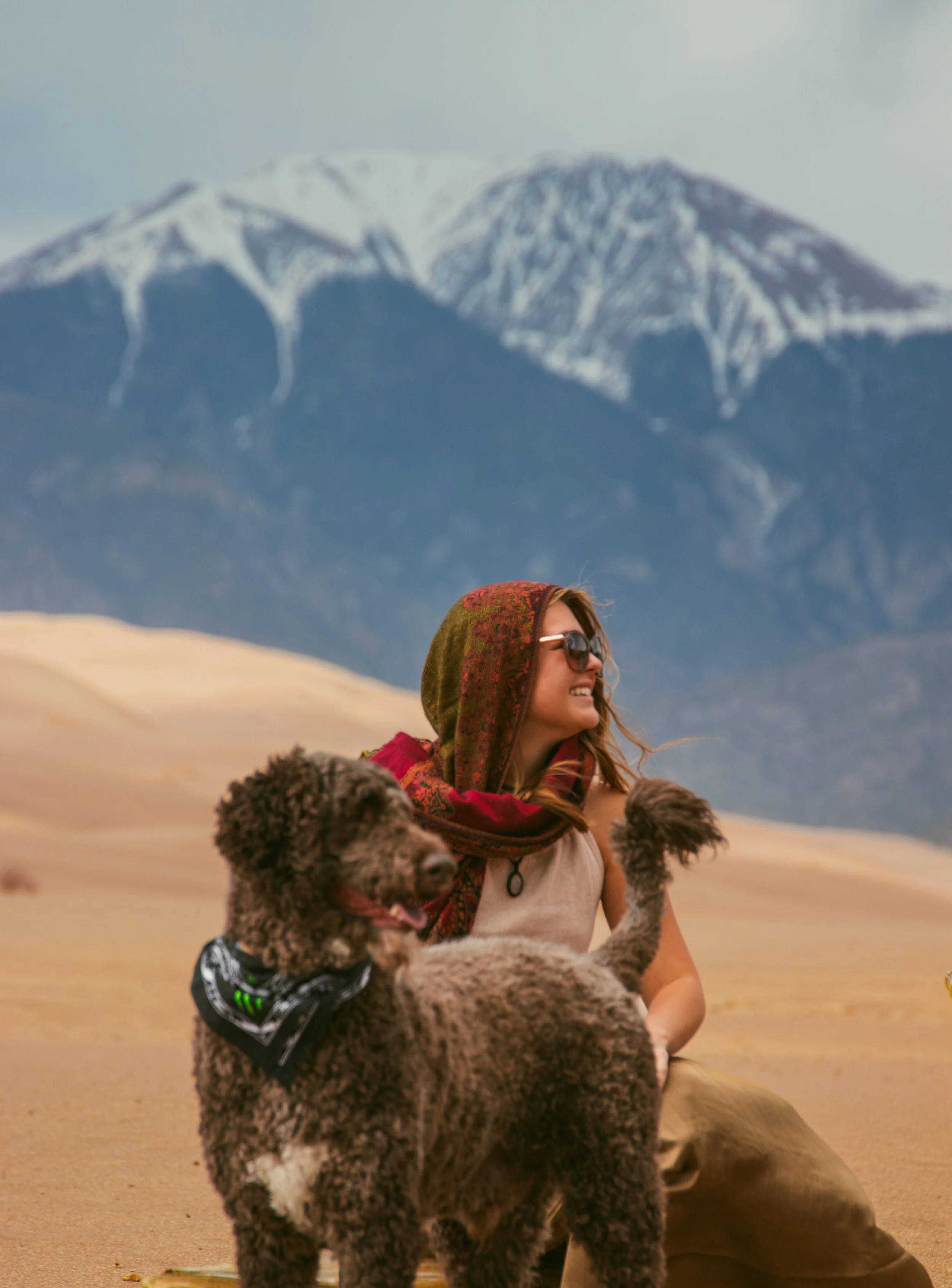 Woman sitting outdoors with a large poodle dog near a mountain landscape. The woman is wearing sunglasses, a colorful scarf, and a sleeveless top. The background features snow-capped mountains and a desert-like terrain.