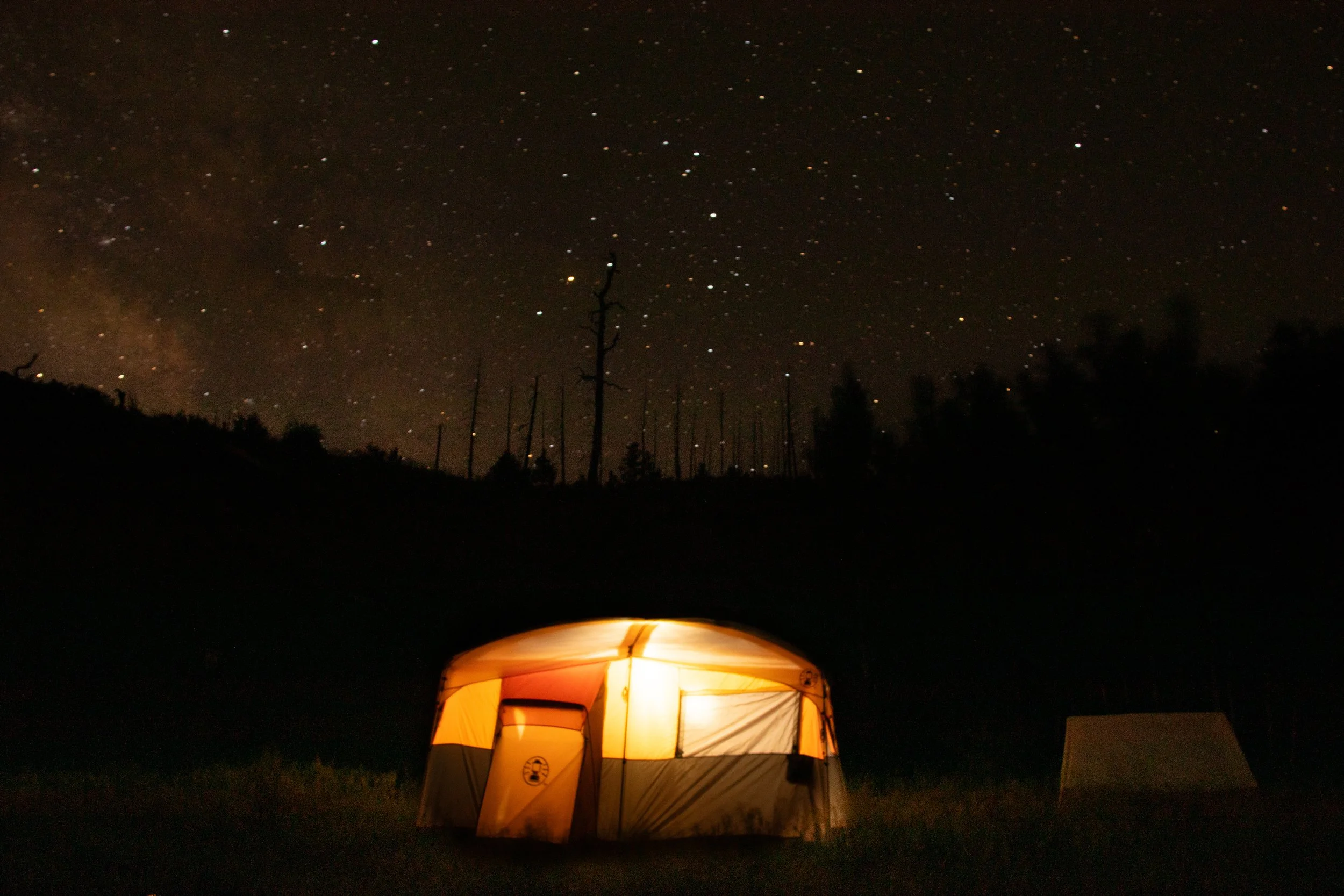 A camping tent illuminated from within under a starry night sky, with silhouettes of trees and pine trees in the background.
