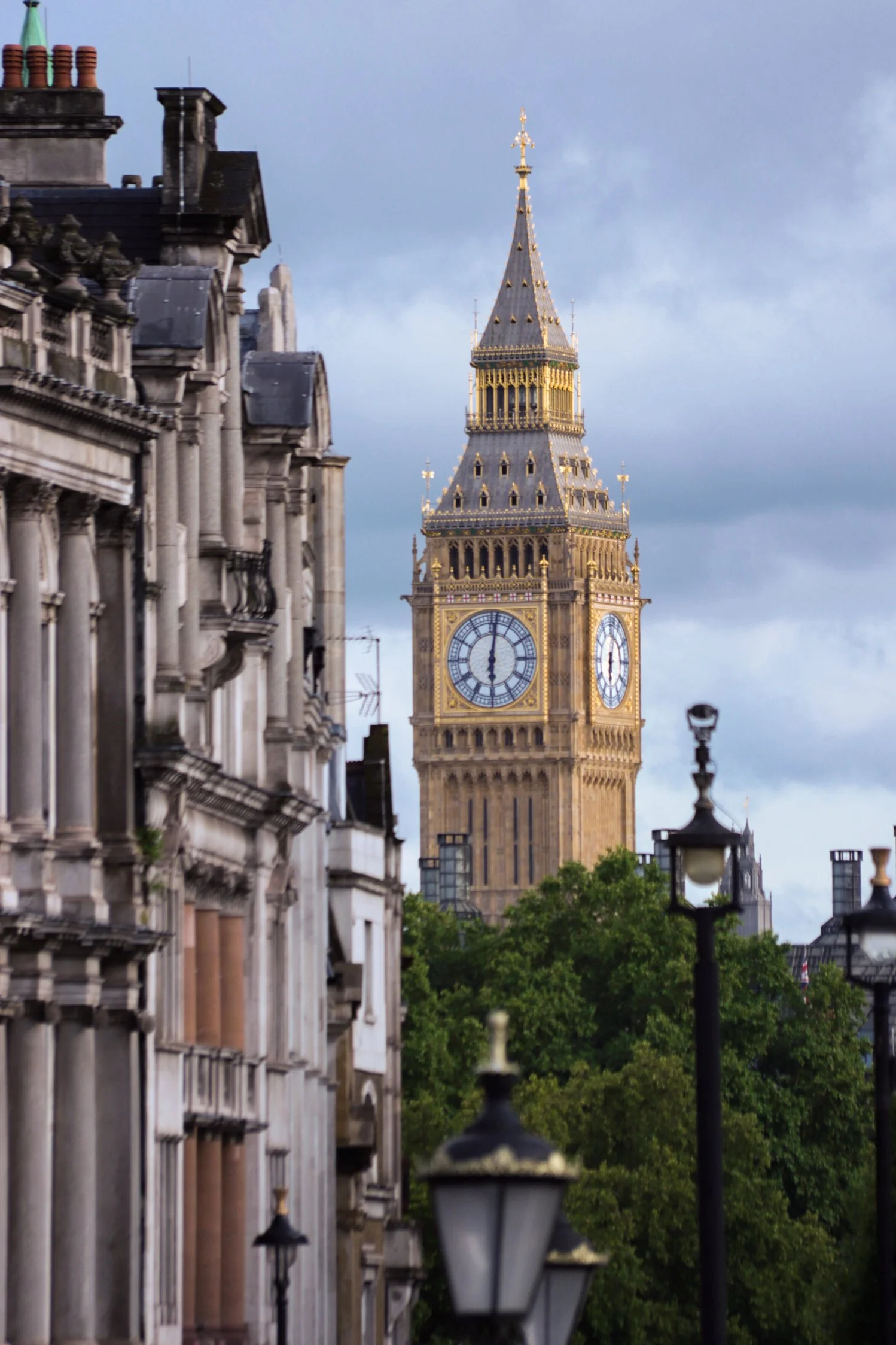 View of Big Ben clock tower in London with historic buildings and street lamps in the foreground, green trees, and a cloudy sky.