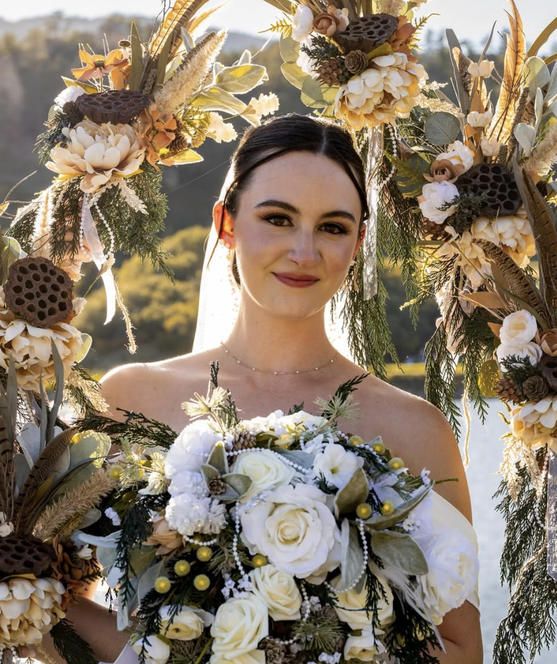 A bride with dark hair and light skin, smiling, stands outdoors during sunset, holding a bouquet of white roses, complemented by greenery and small yellow accents. She is framed by an arch decorated with flowers, leaves, and dried natural elements, with a scenic background of hills and a lake.
