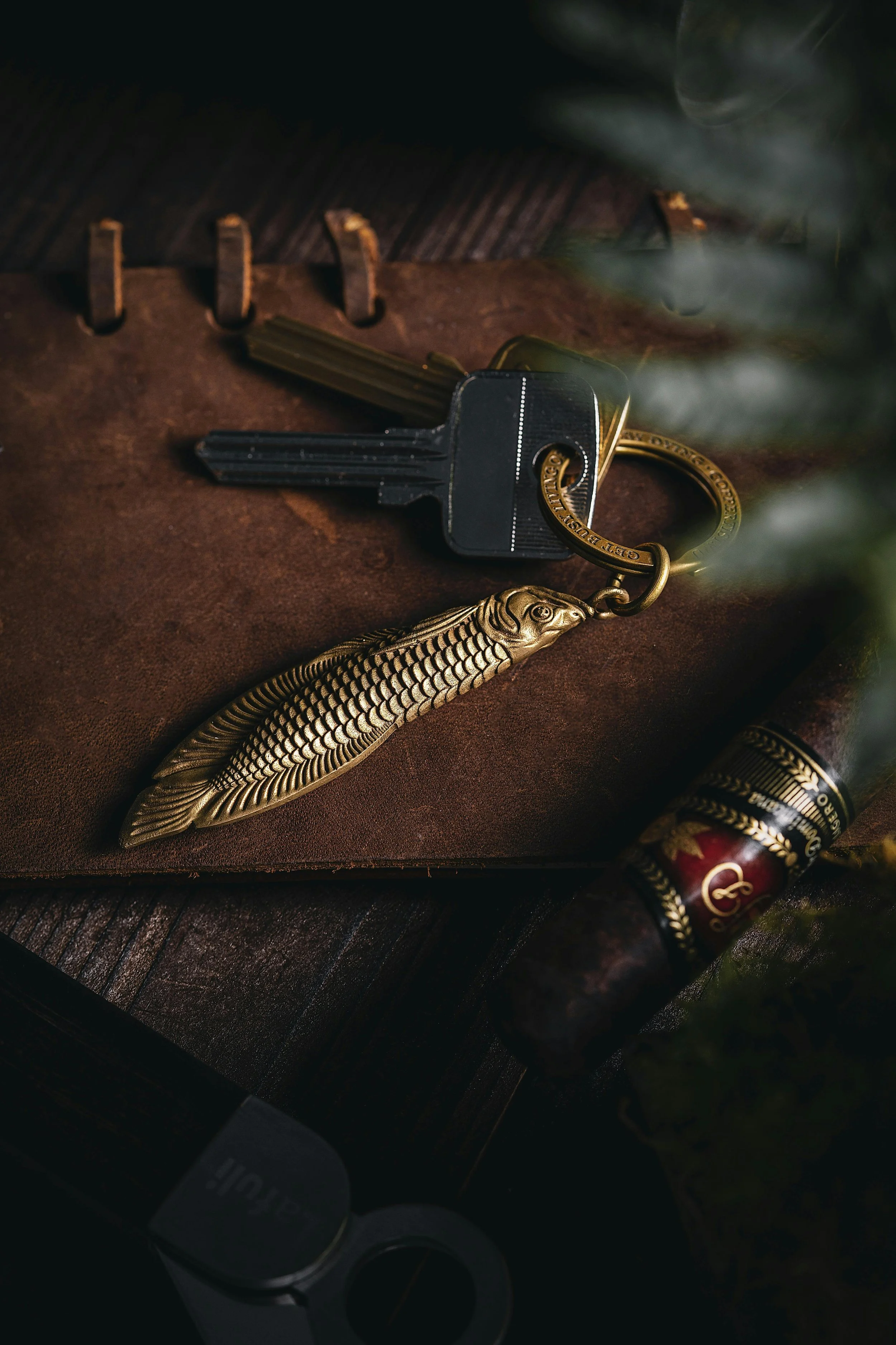 Close-up of a set of keys with a decorative fish-shaped keychain, a cigar, and some greenery on a leather surface.