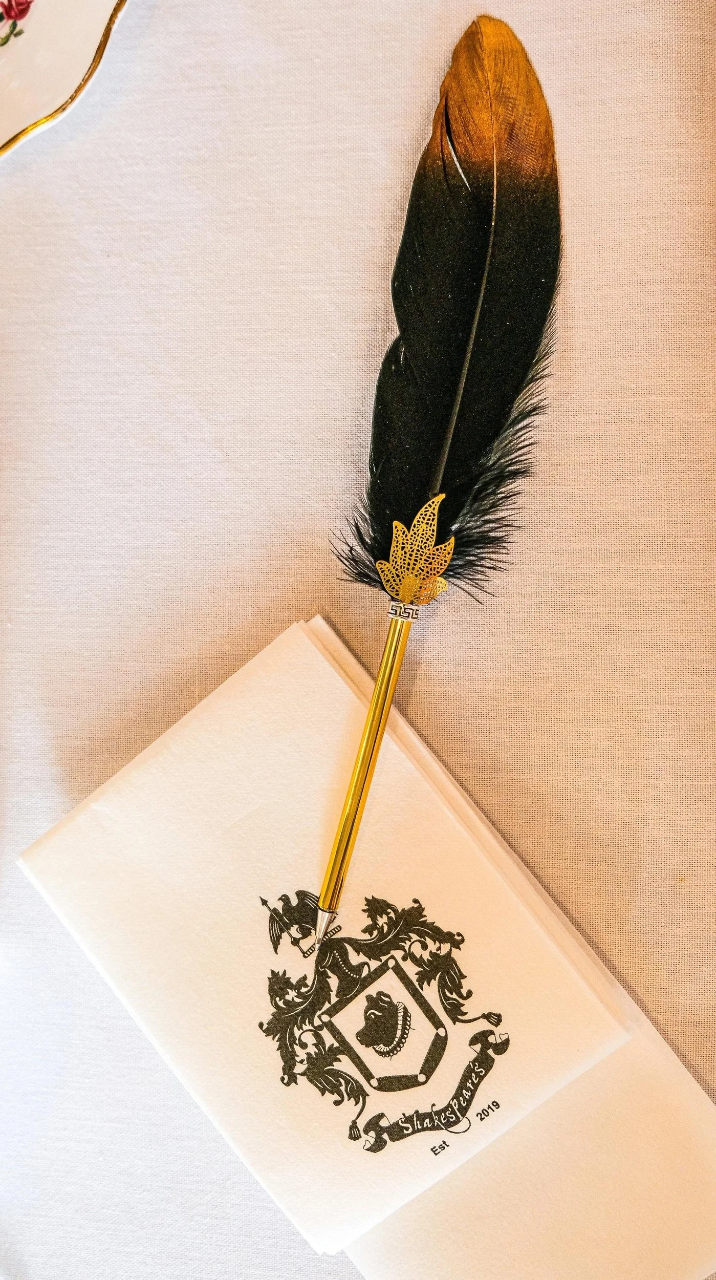 A gold and black feather quill pen with a decorative gold leaf design at its base, resting on a white paper with a black Shakespeares crest and establishment date of 2019.