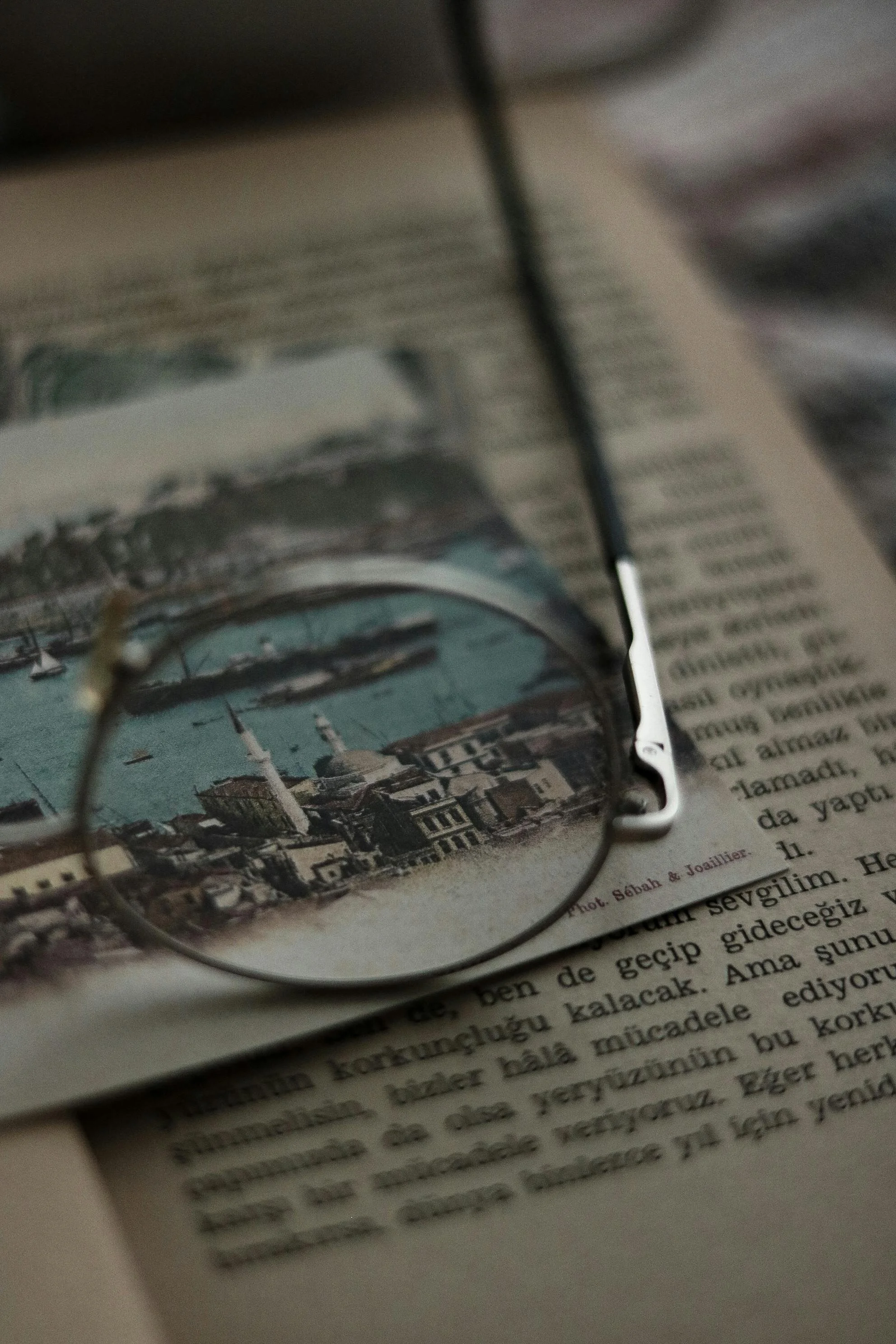 A magnifying glass on top of a page from a book, focusing on a colored illustration of a coastal city with mosques, water, and boats.