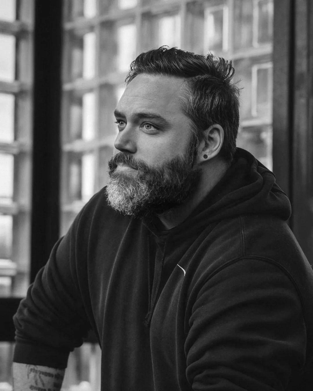 A black and white photo of a bearded man with a hairstyle, wearing a hoodie, sitting indoors near a window, looking contemplative.