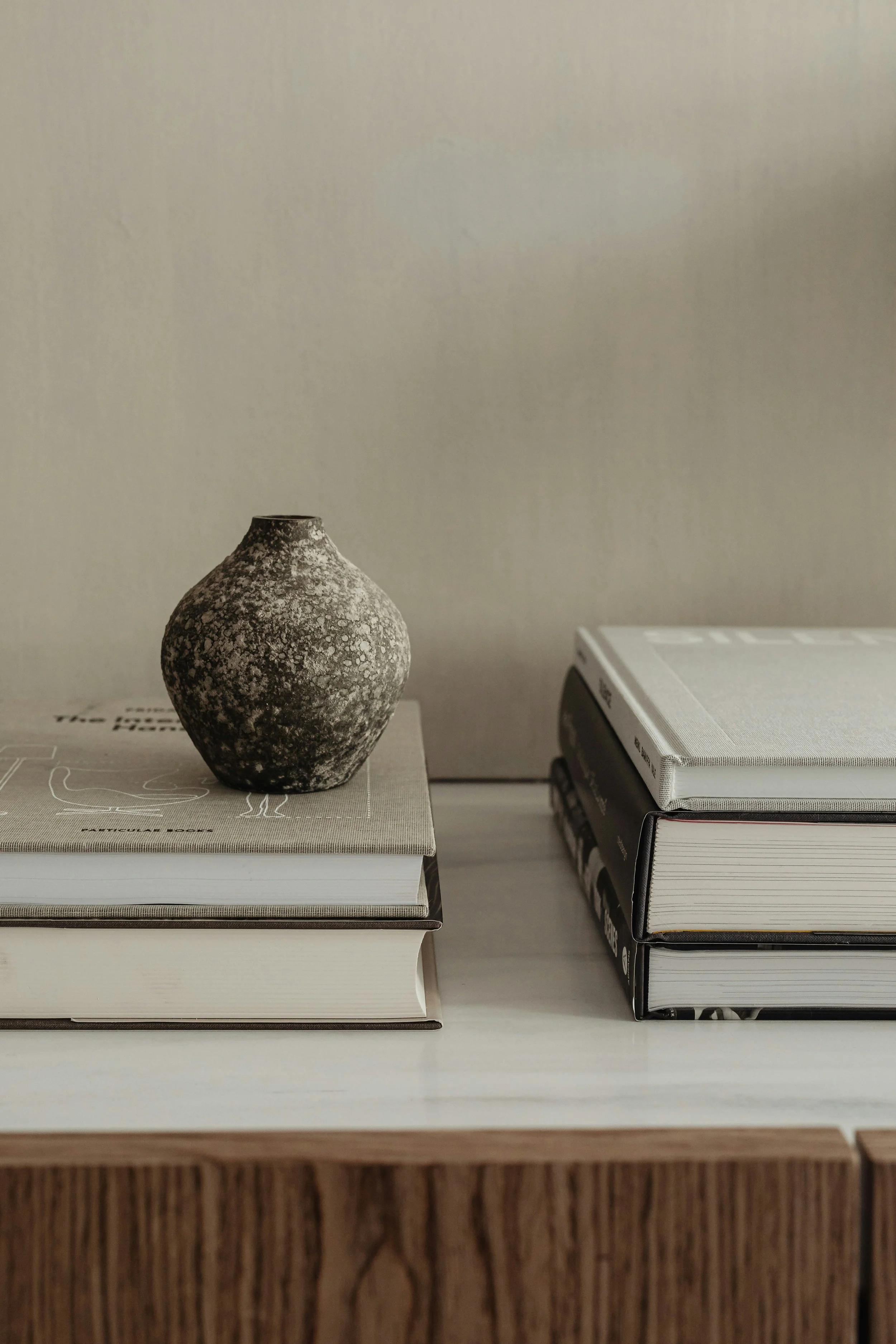 A small, textured dark-brown and gray ceramic vase sitting on top of three stacked books on a wooden surface with a plain beige wall in the background.