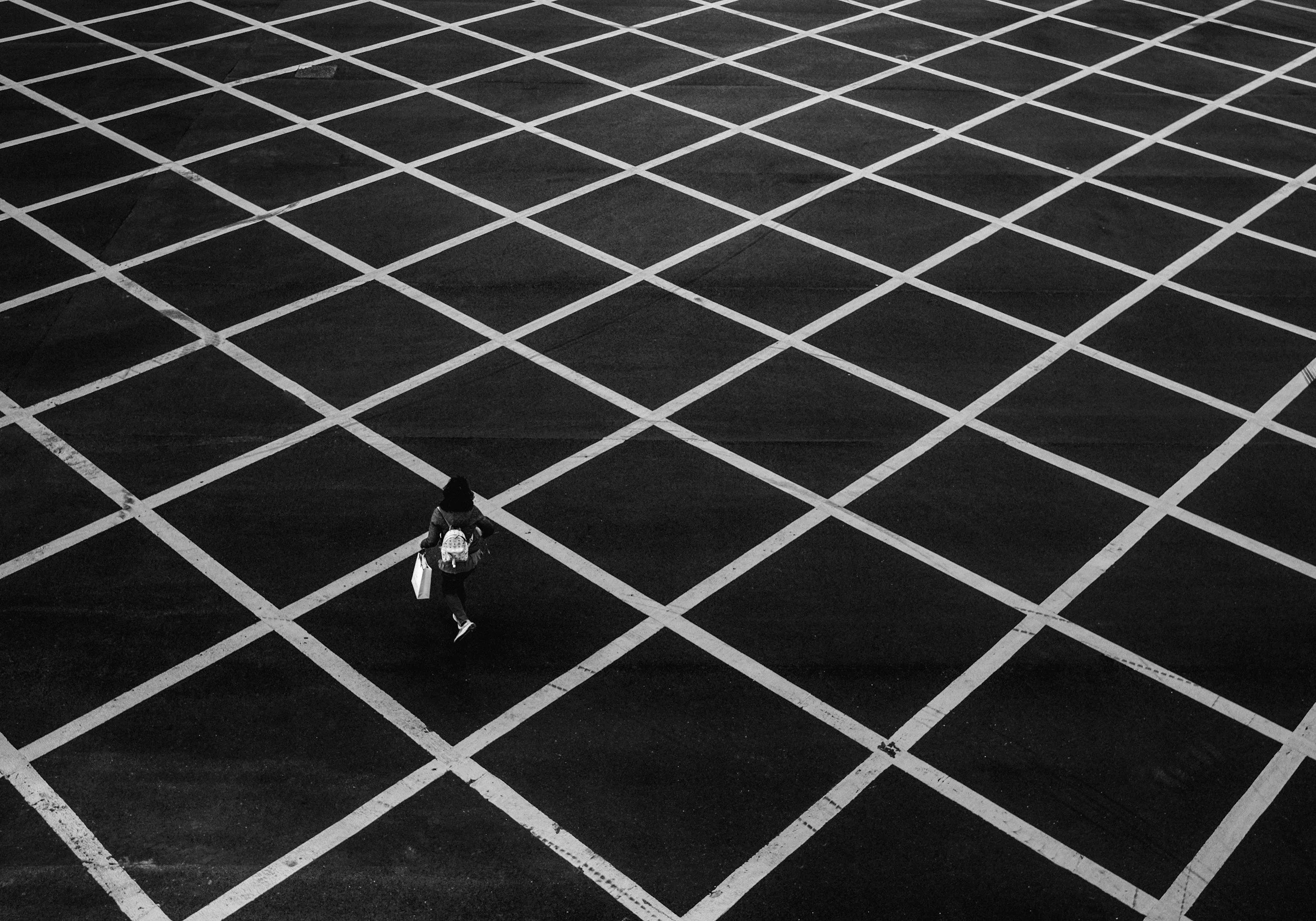 An overhead view of a mostly empty parking lot with a single person walking across the marked spaces, carrying shopping bags.