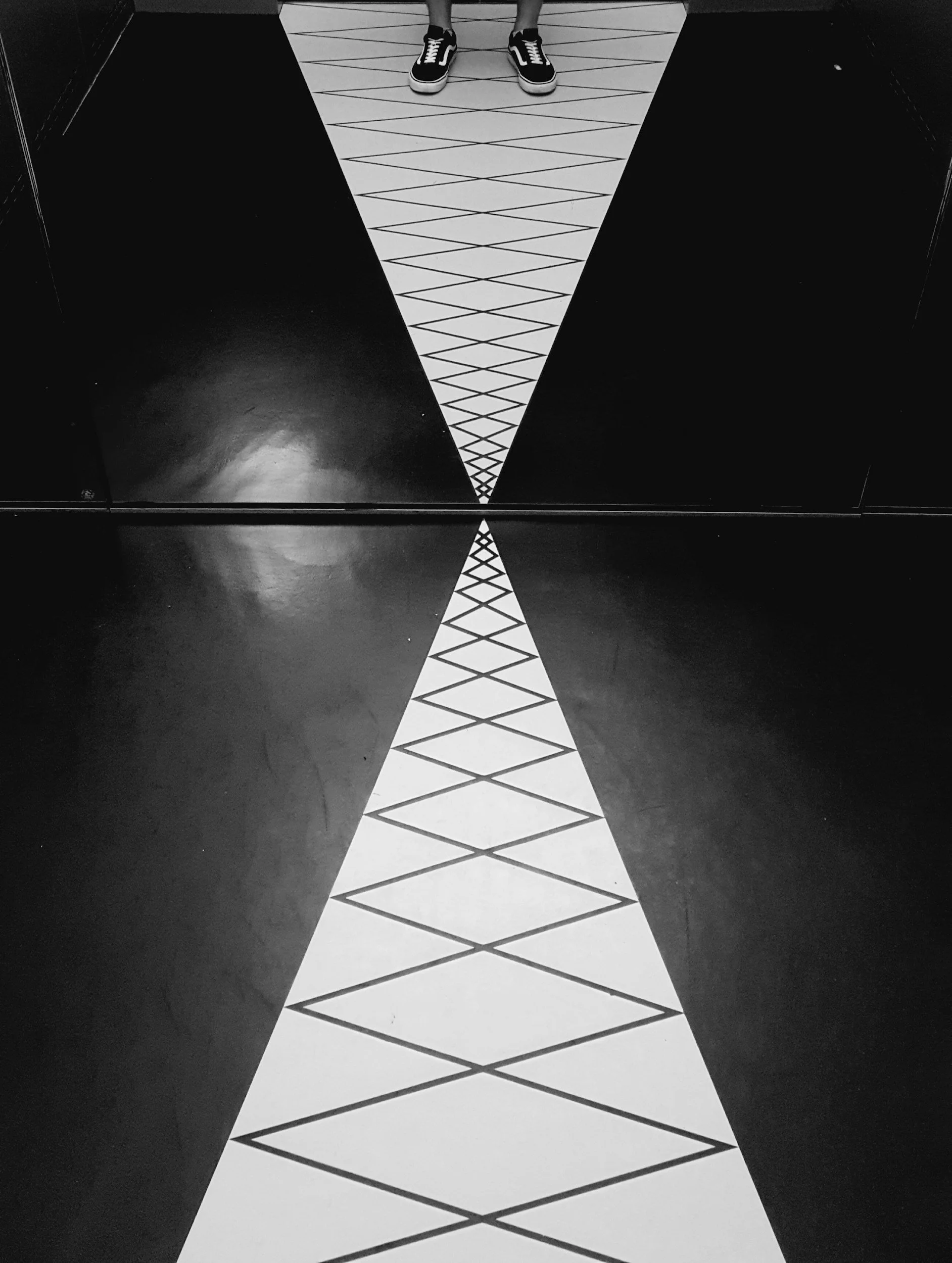 A black and white photo of a person standing in front of a mirror, with only their legs visible. The mirror has a triangular design with a geometric pattern leading to a point at the bottom.