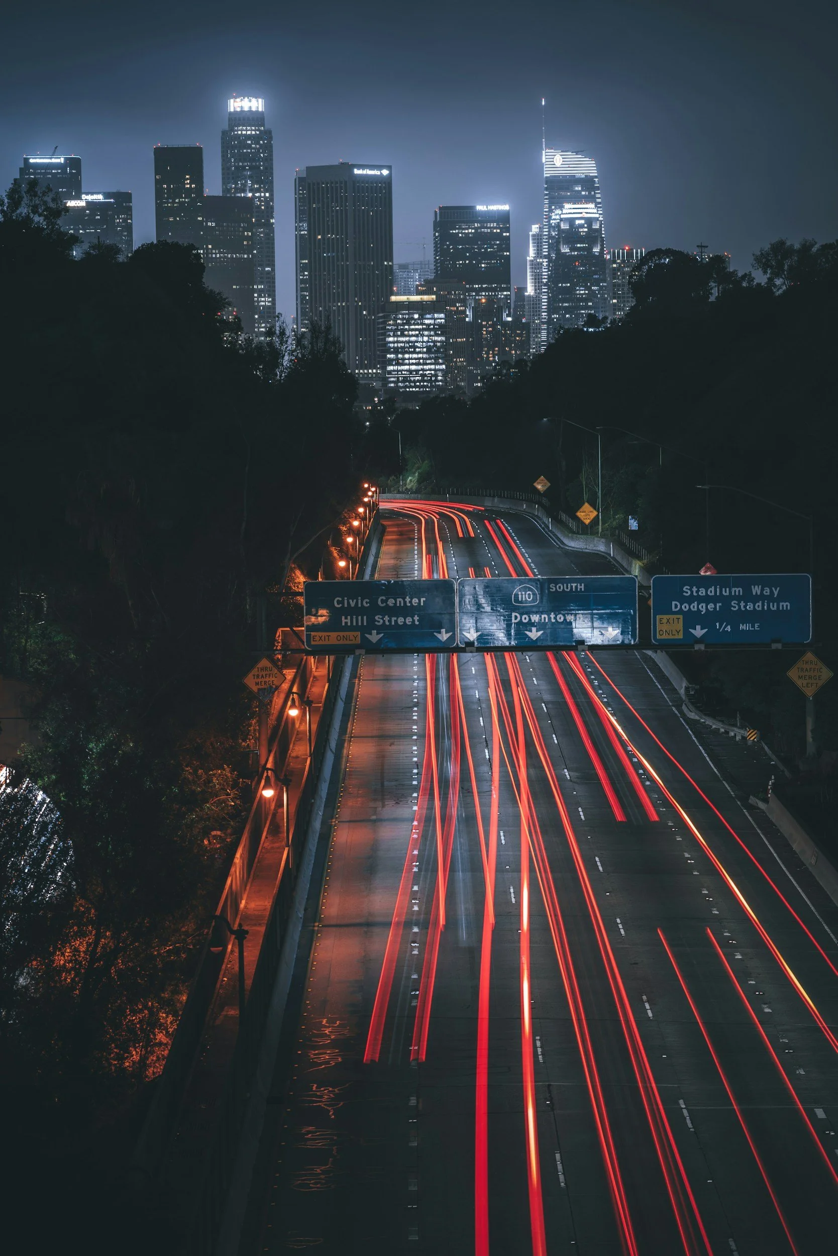 Nighttime city skyline with tall illuminated skyscrapers and busy highway with red light trails from moving vehicles.