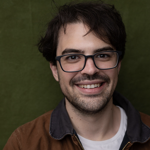 A young man with dark hair, glasses, and a beard smiling at the camera against a green background.