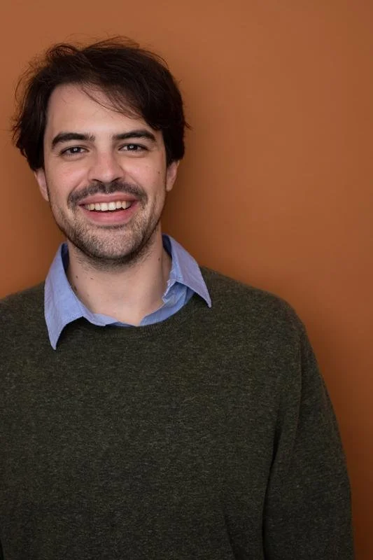 Portrait of a smiling young man with dark hair and facial hair, wearing a blue collared shirt under a dark sweater, standing against a brown background.