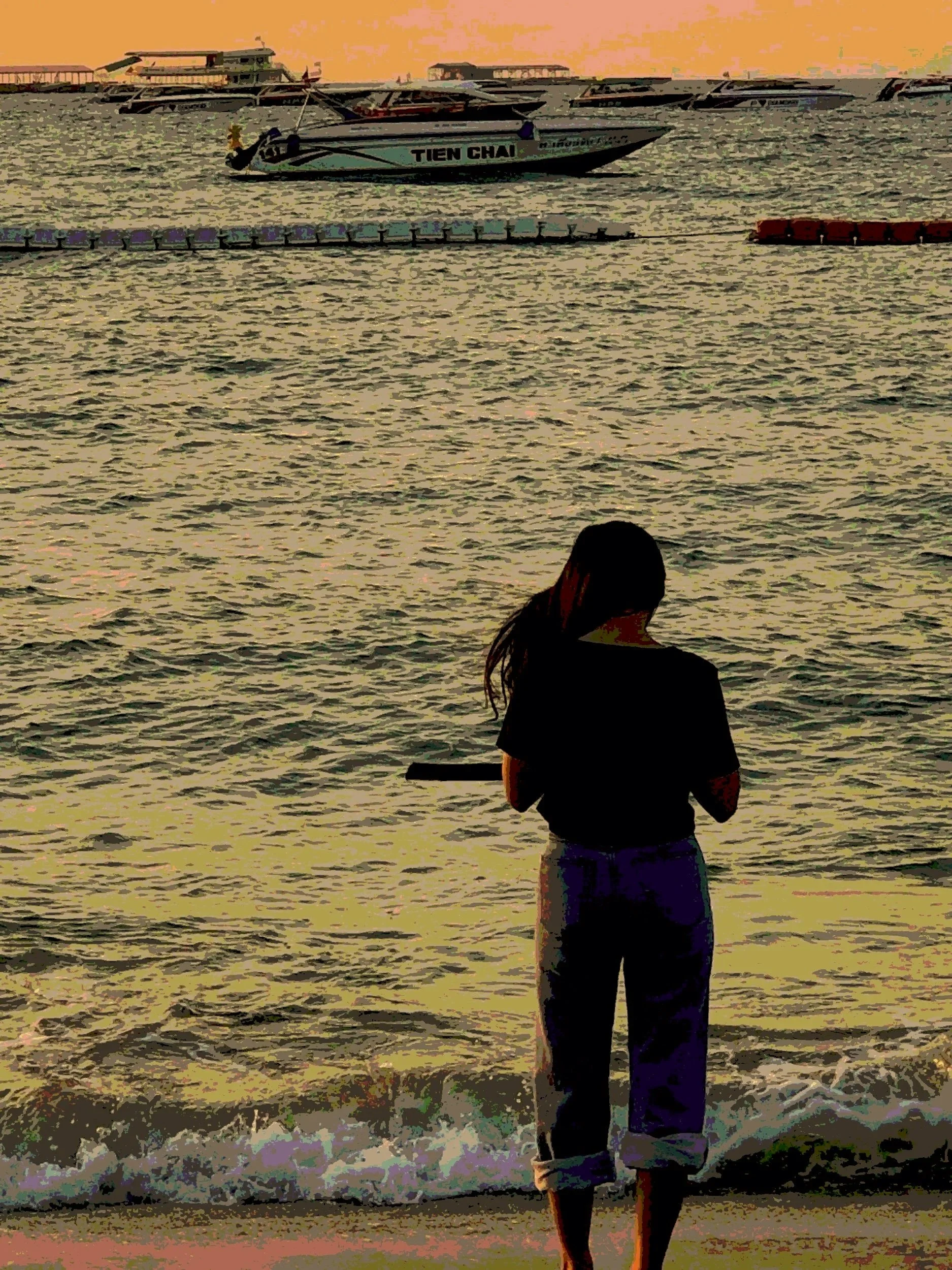 A woman standing at the shoreline, facing the water at sunset with boats and a pier in the background.