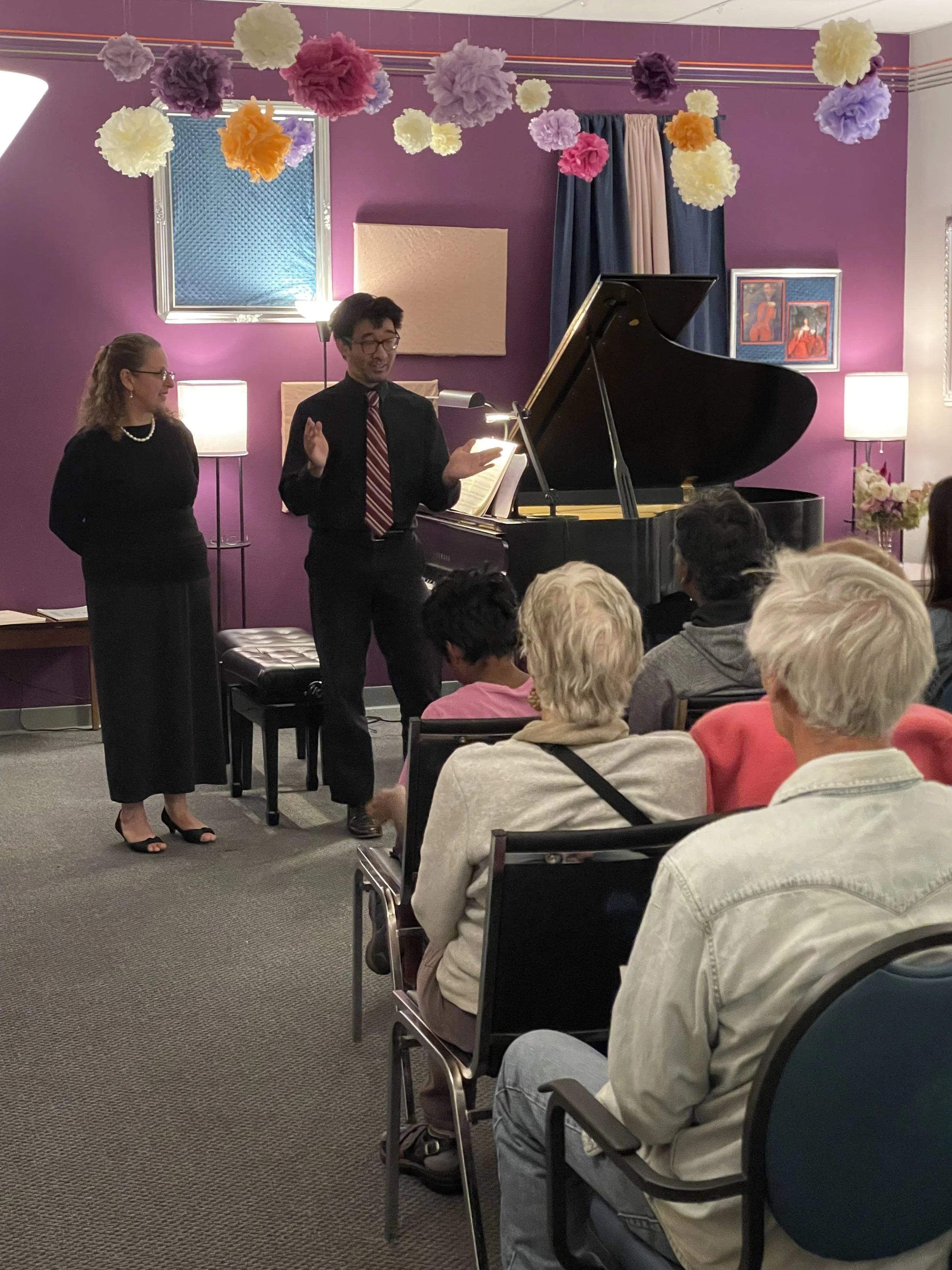 A man and a woman are standing near a grand piano in a room decorated with colorful paper pom-poms hanging from the ceiling. The man appears to be speaking or performing, with an open book or music sheet in front of him, while the woman looks on smiling. An audience is seated, watching the performance in a cozy, warmly-lit room with purple walls.