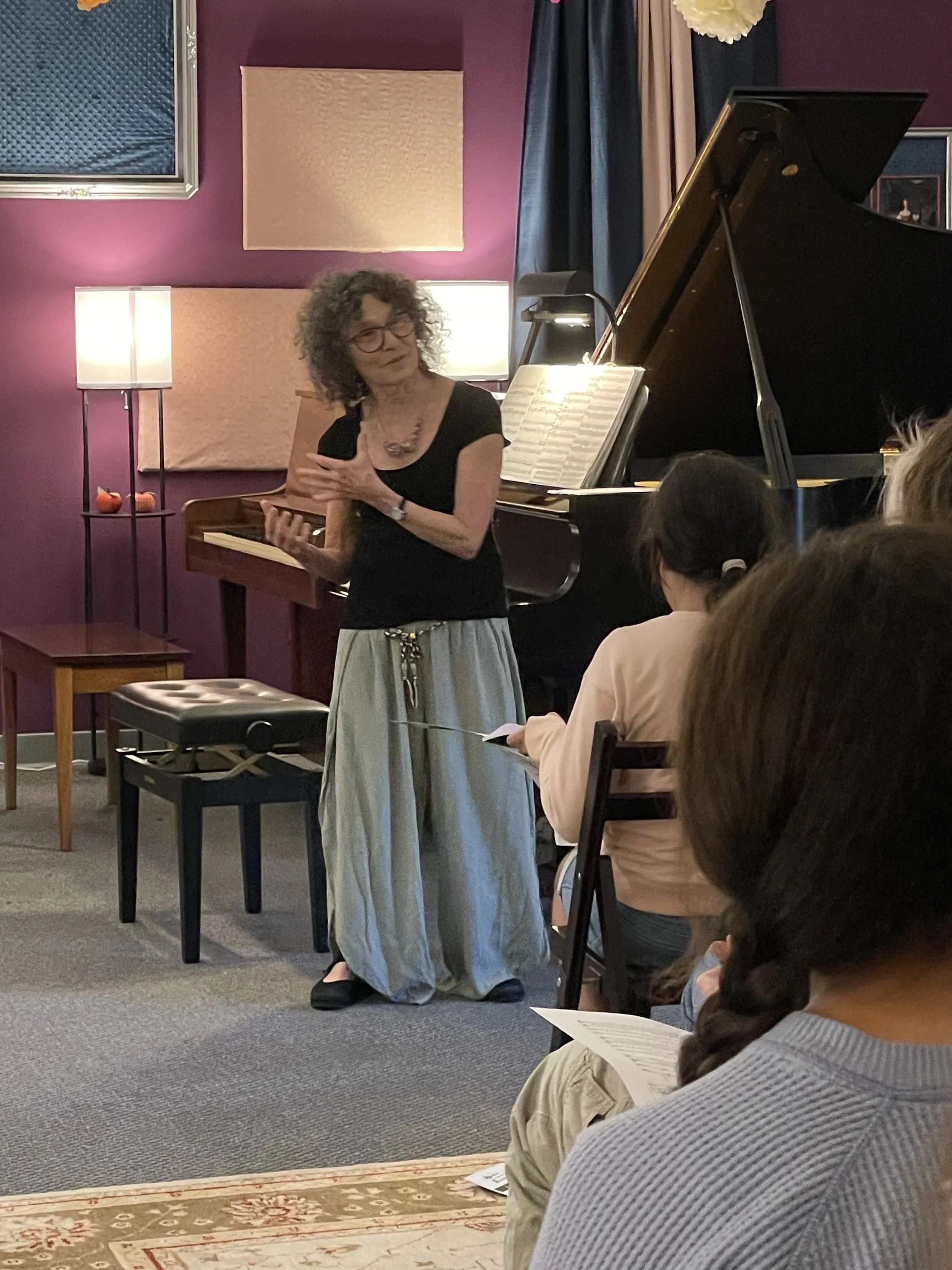 A woman with curly hair and glasses is standing in front of a piano, speaking to an audience in a room with purple walls, acoustic panels, and a table lamp. She is wearing a black top and a long, flowing skirt, and is gesturing with her hands while holding a piece of paper. Some audience members are seated, holding papers or notebooks, with one person in the foreground having a braid.