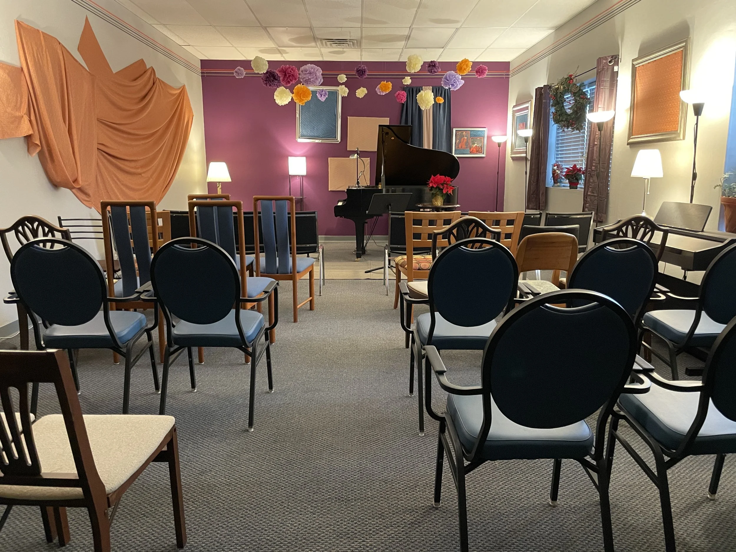 Colorful and cozy recital space with a grand piano, purple accent wall, hanging paper flowers, and rows of chairs facing the piano.