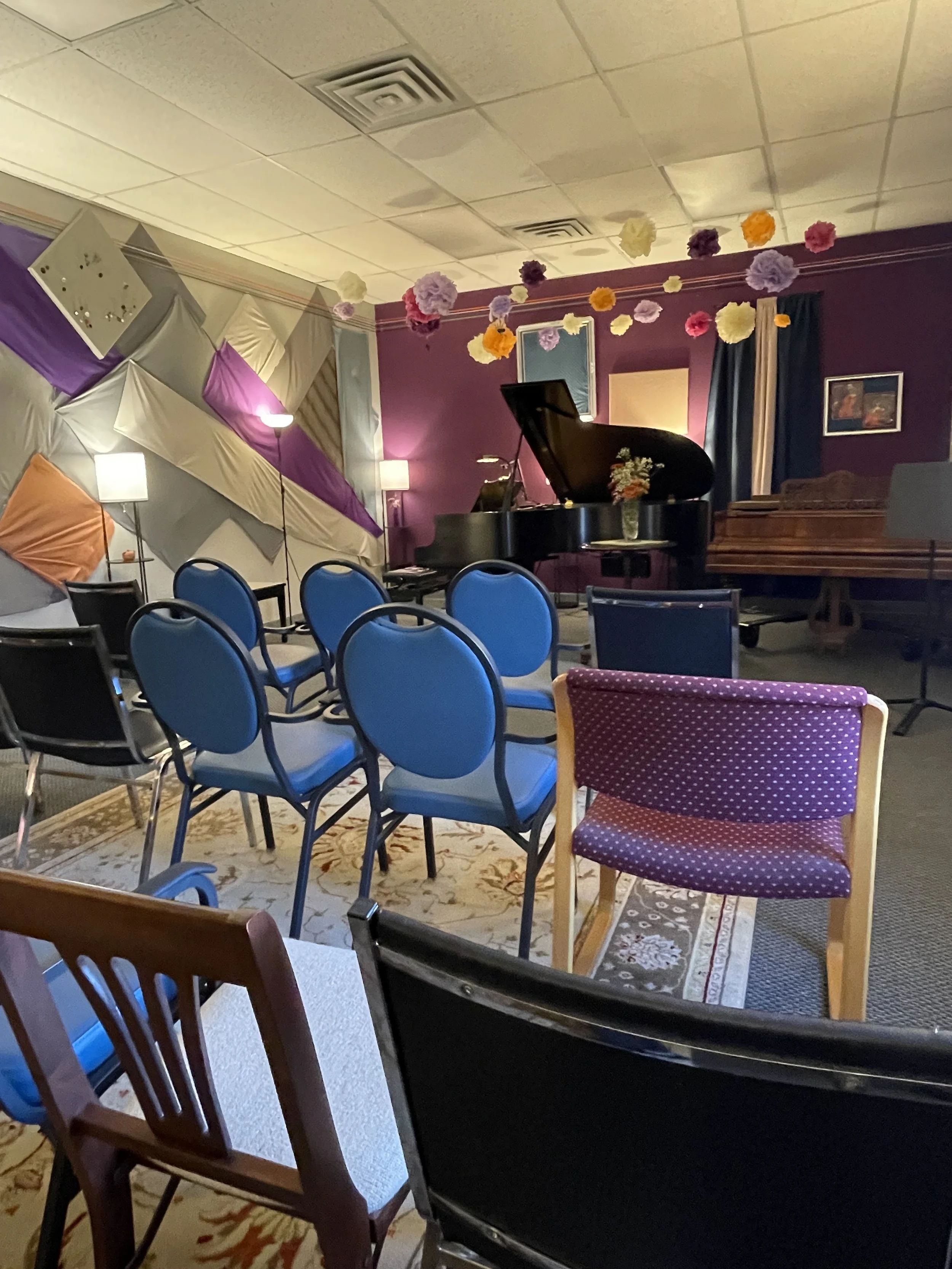 A small performance space with a grand piano, colorful hanging paper flowers decorate the space. Chairs are arranged for an audience, making this the perfect space for your next recital.
