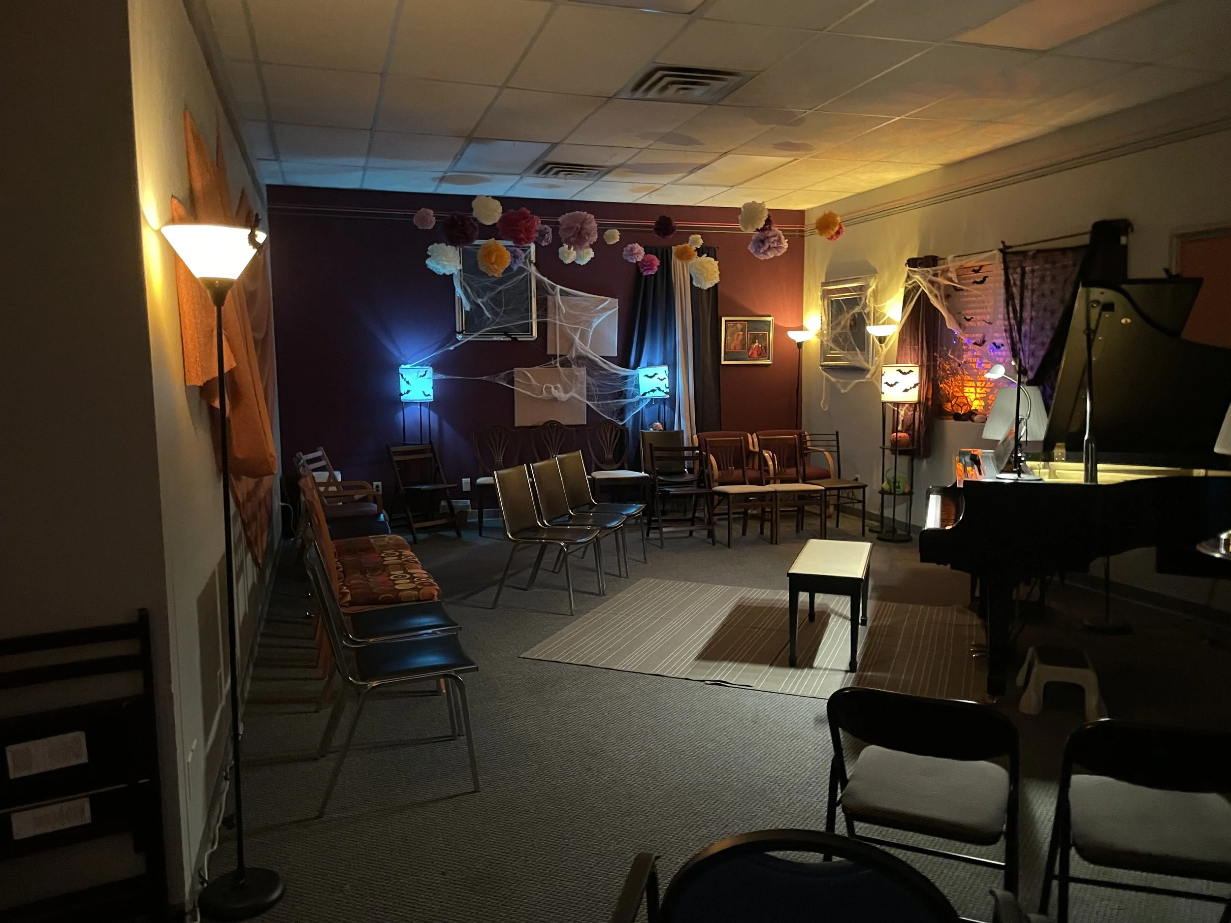 A decorated room with Halloween-themed decorations, including spider webs, paper flowers, and jack-o'-lantern patterned lamps. There are chairs arranged along the walls, a small table on a striped rug, and a grand piano on the right. The room is dimly lit with warm and colorful lighting.
