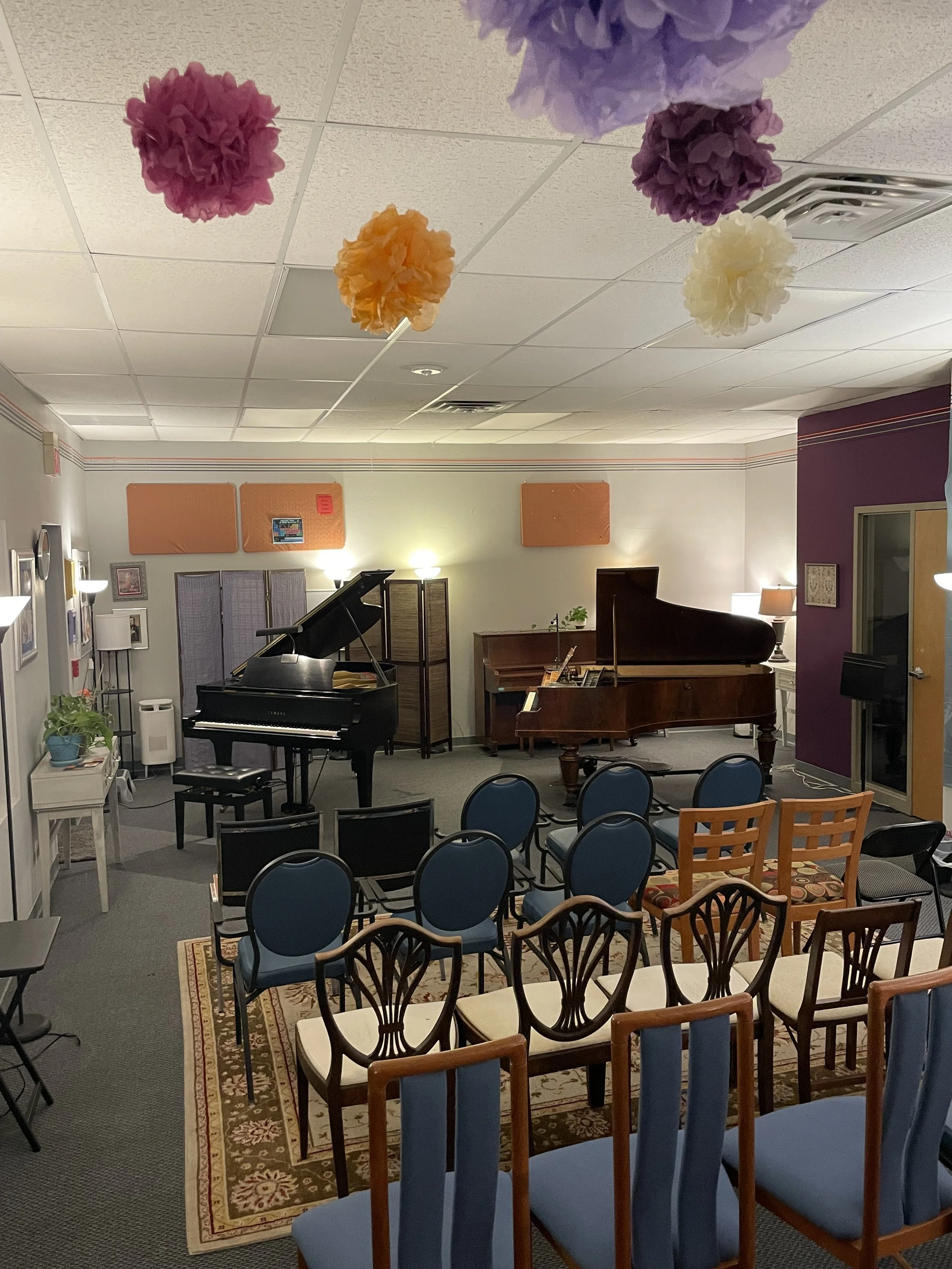 Room decorated with colorful tissue paper pom-poms hanging from the ceiling, arranged for a musical or performance event, with chairs facing towards two pianos and a small stage area.