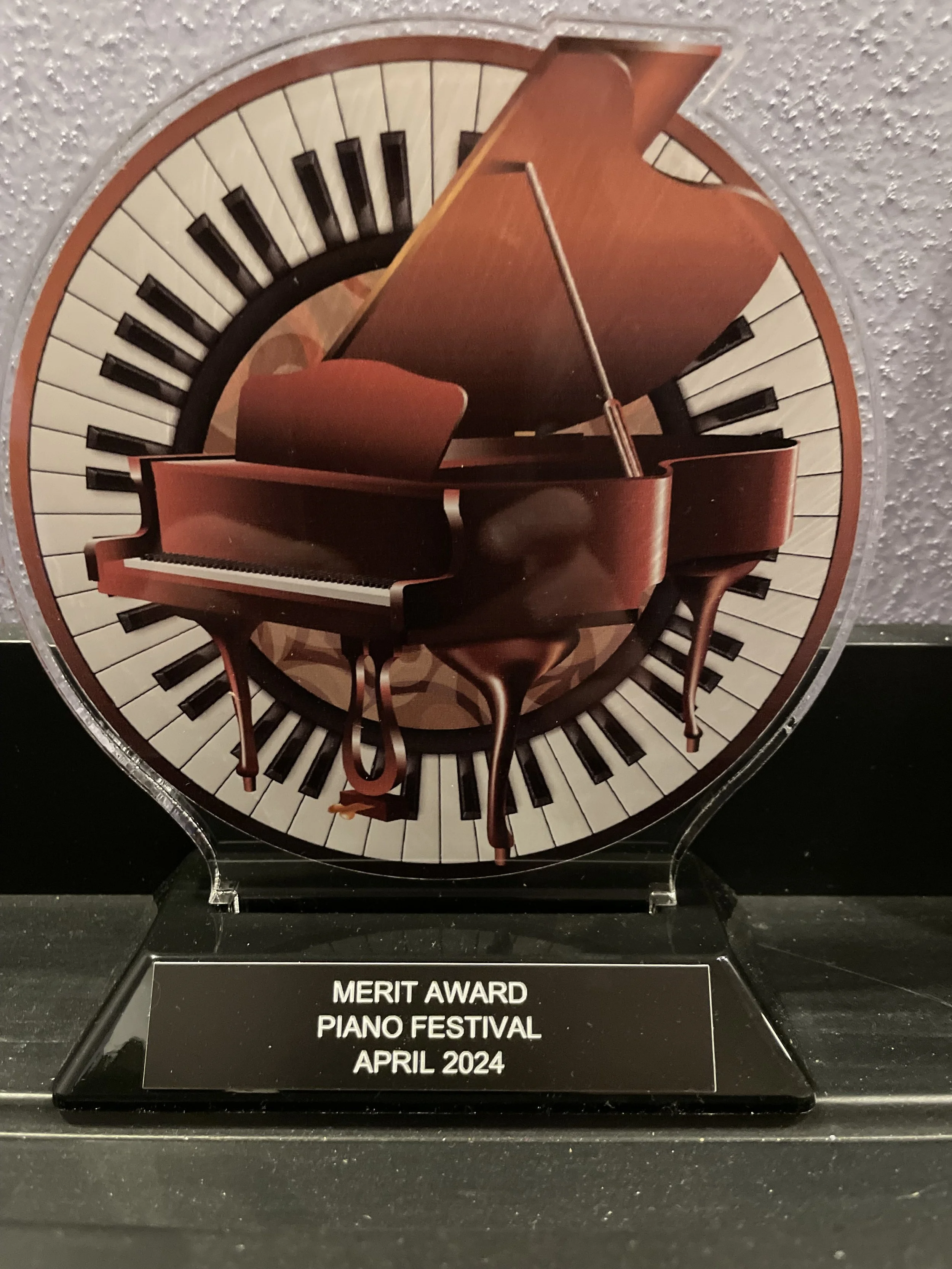 Trophy with a piano and piano keys design, inscribed with "MERIT AWARD PIANO FESTIVAL APRIL 2024."