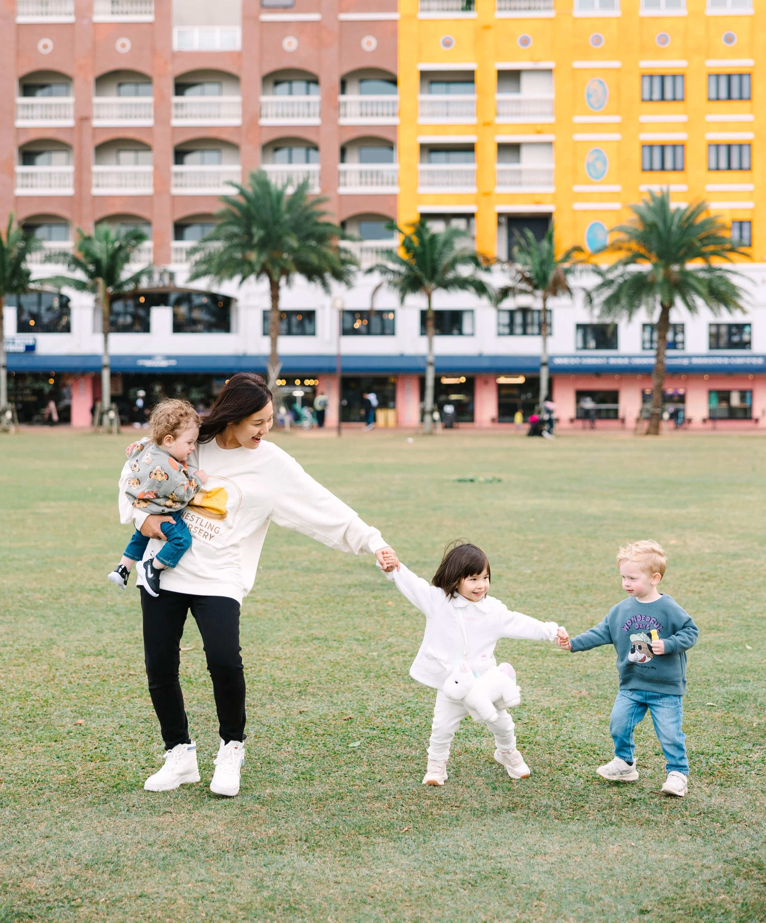 A woman playing with two young international children in a park, holding the hands of a girl and a boy, with a colorful building and palm trees in the background.