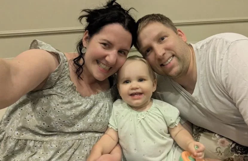 A woman, a man, and a toddler taking a selfie together. The woman has dark hair and is smiling, the man has short hair and is smiling, and the toddler is sitting between them, smiling and holding a pacifier.