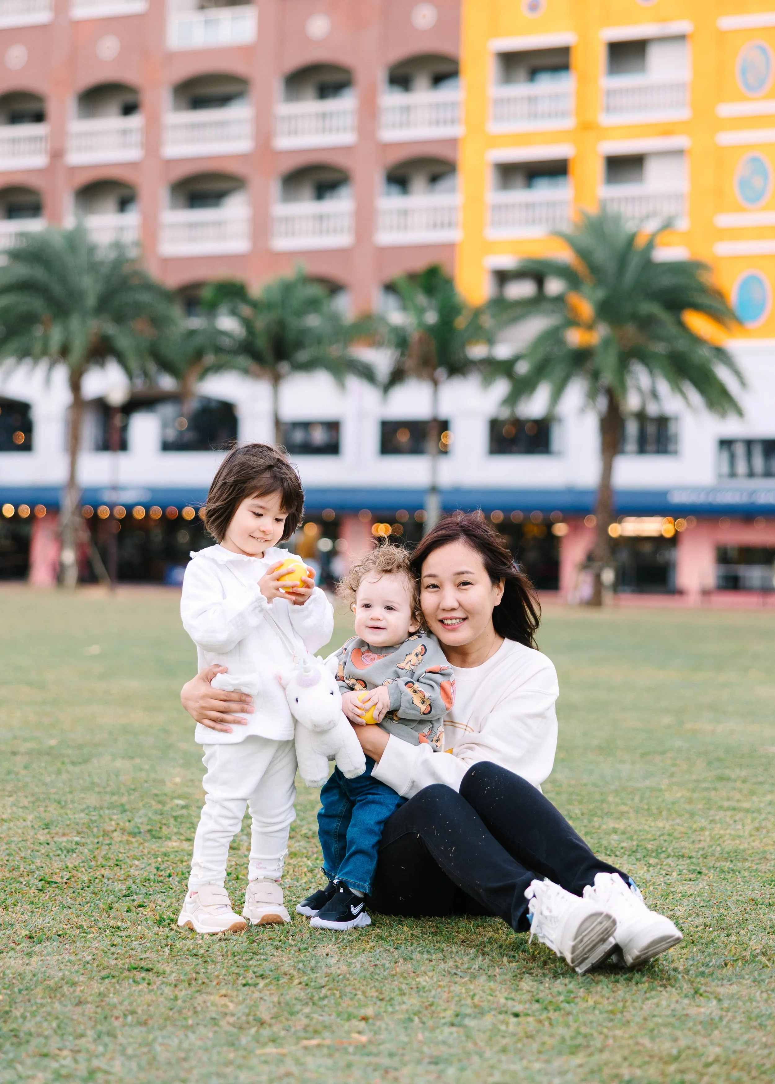 A woman sitting on the grass with two children, one holding a toy unicorn, in front of a hotel or apartment building with palm trees.