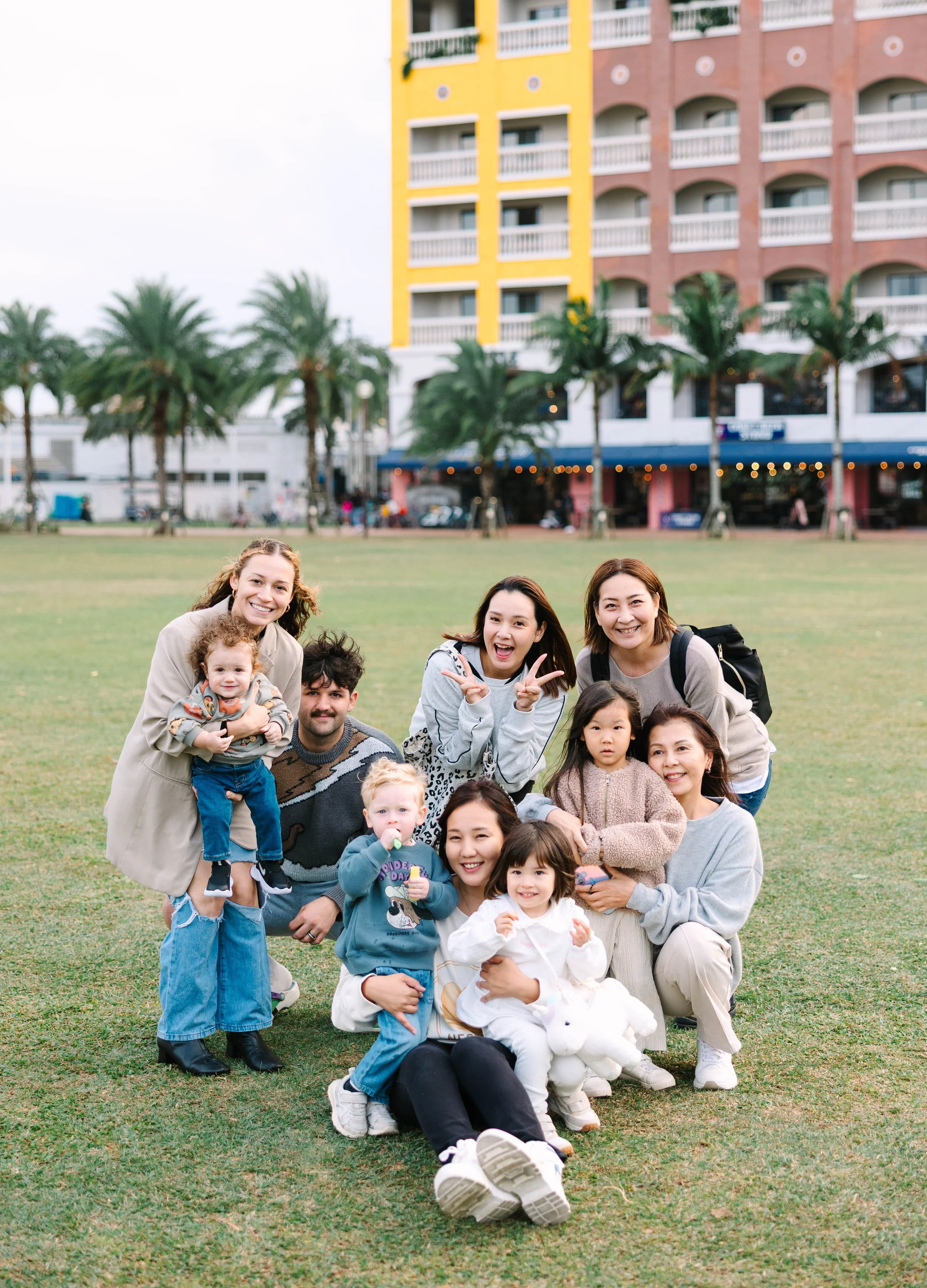 Group of ten people, including children and adults, posing outdoors on a grassy field in front of a building with palm trees. Everyone is smiling and making playful gestures.