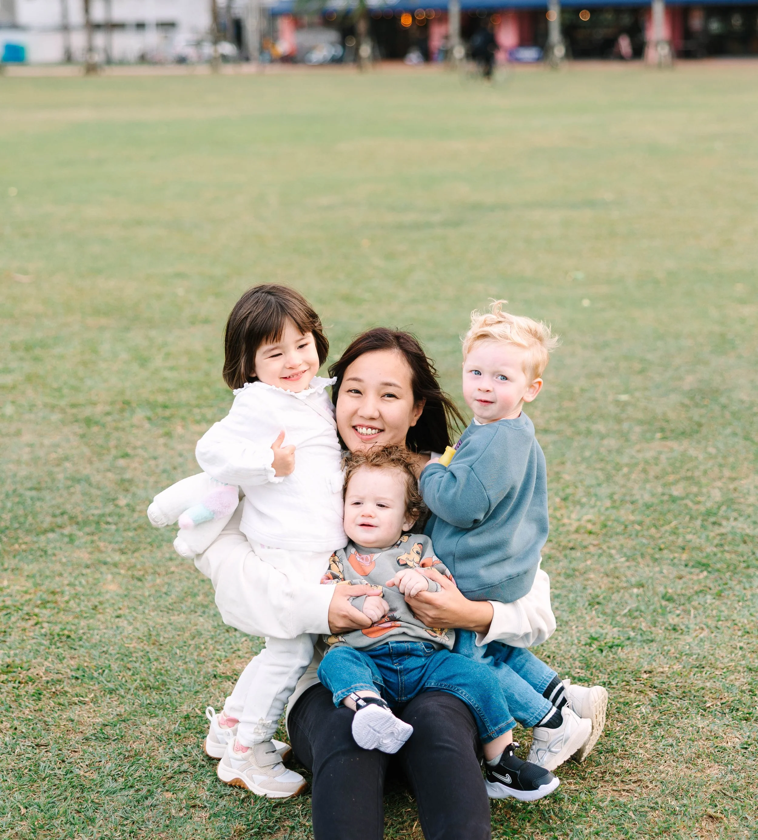 A woman sitting on the grass holding three young children, smiling and posing for the camera at a park.