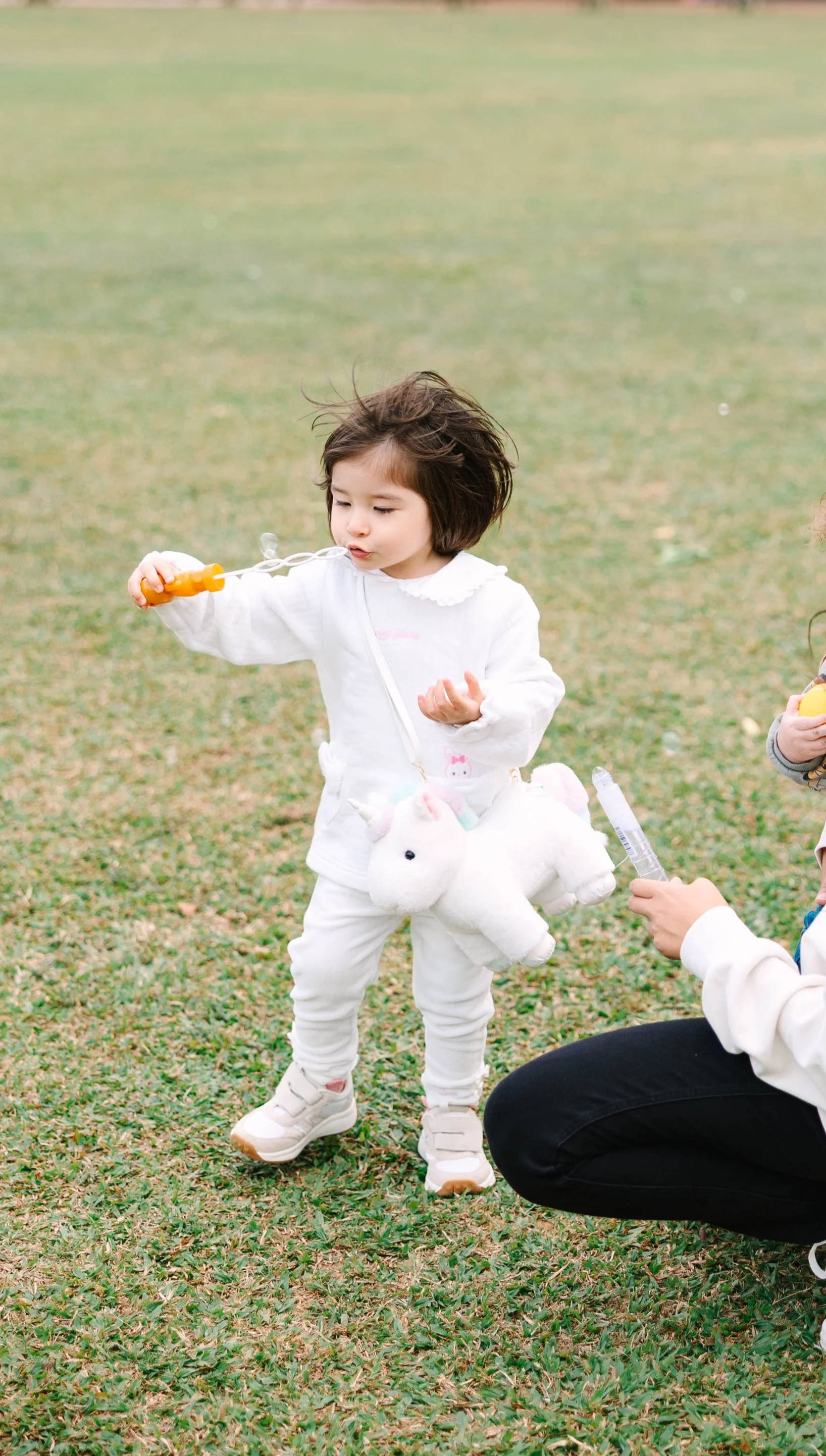 Young girl with a unicorn plush bag blowing bubbles outdoors on the grass, with another child holding bubble toys nearby.