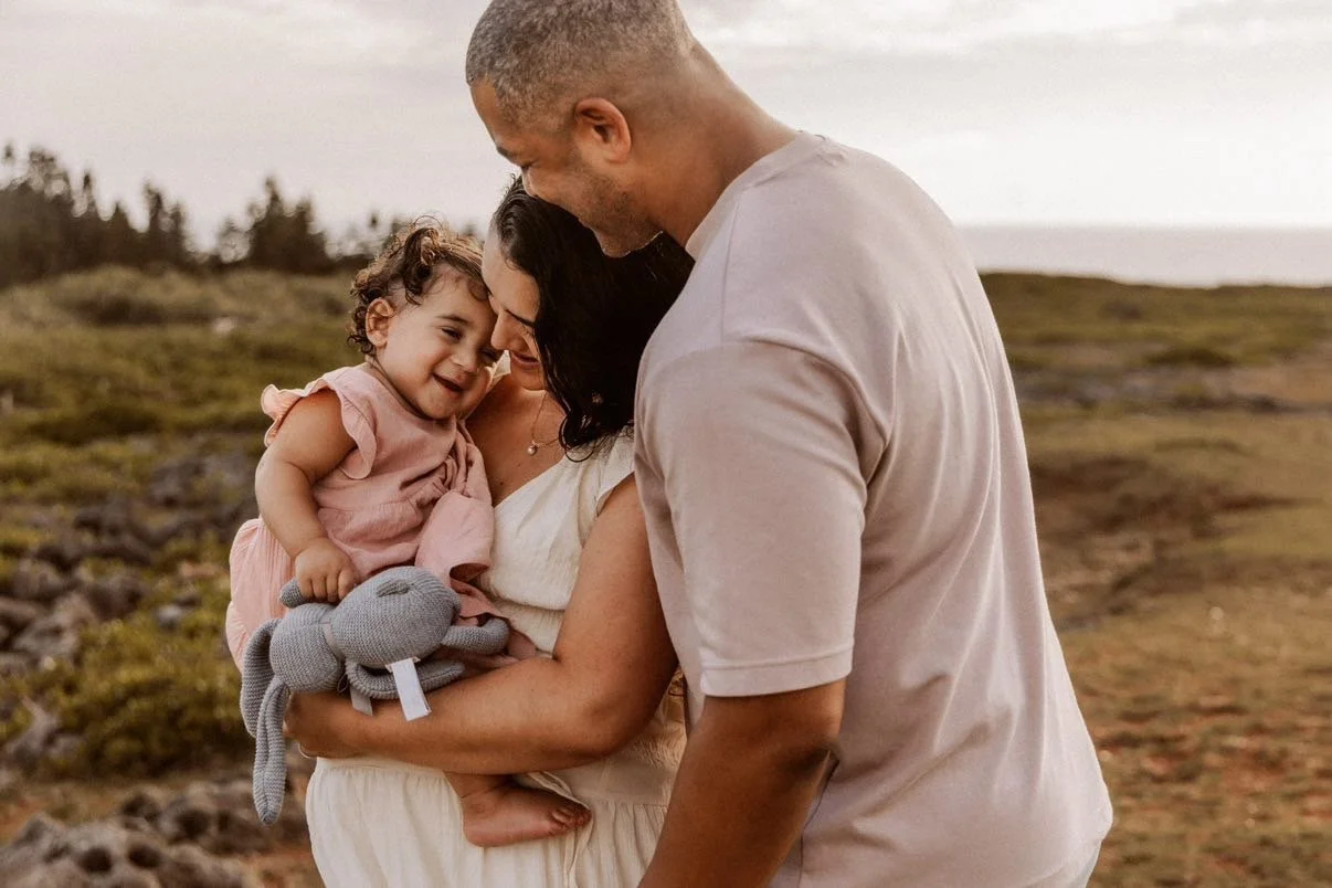 A family of three, a woman, a man, and a young girl, standing outdoors on a grassy terrain with trees in the background. The woman is holding the girl and a gray stuffed animal, and they are all touching foreheads, sharing a tender moment.