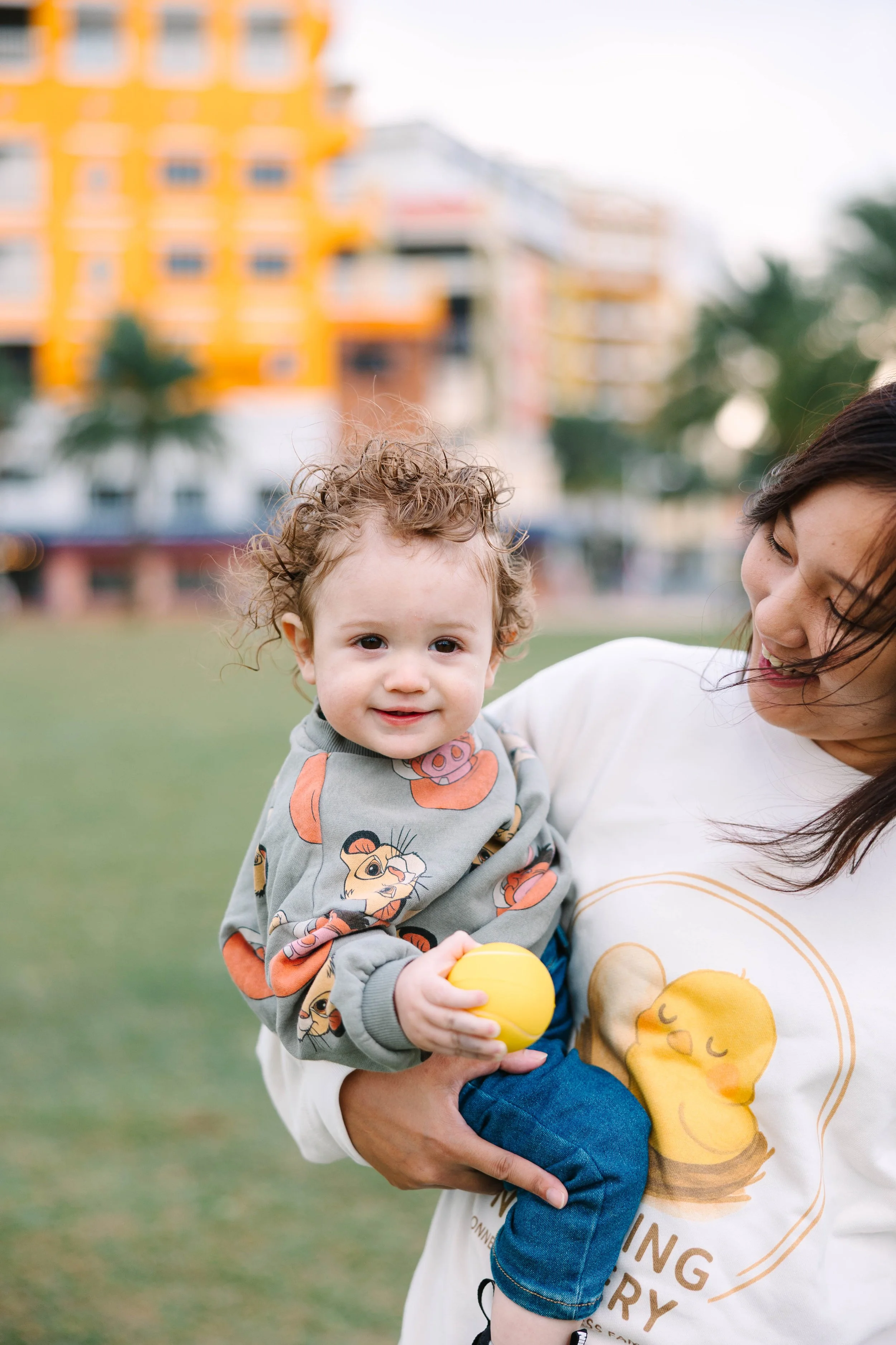 A young child with curly hair smiling while being held by a woman outdoors, with a blurred colorful building in the background.