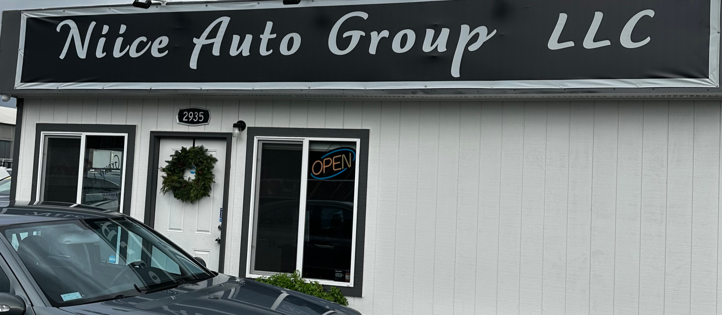 The image shows the storefront of Nice Auto Group LLC with a black sign displaying the business name in white cursive text. There is a wreath hanging on the white door, which also has an orange and blue neon 'OPEN' sign in the window. A parking lot with a silver car is in front of the building, and the building's address 2935 is above the door.